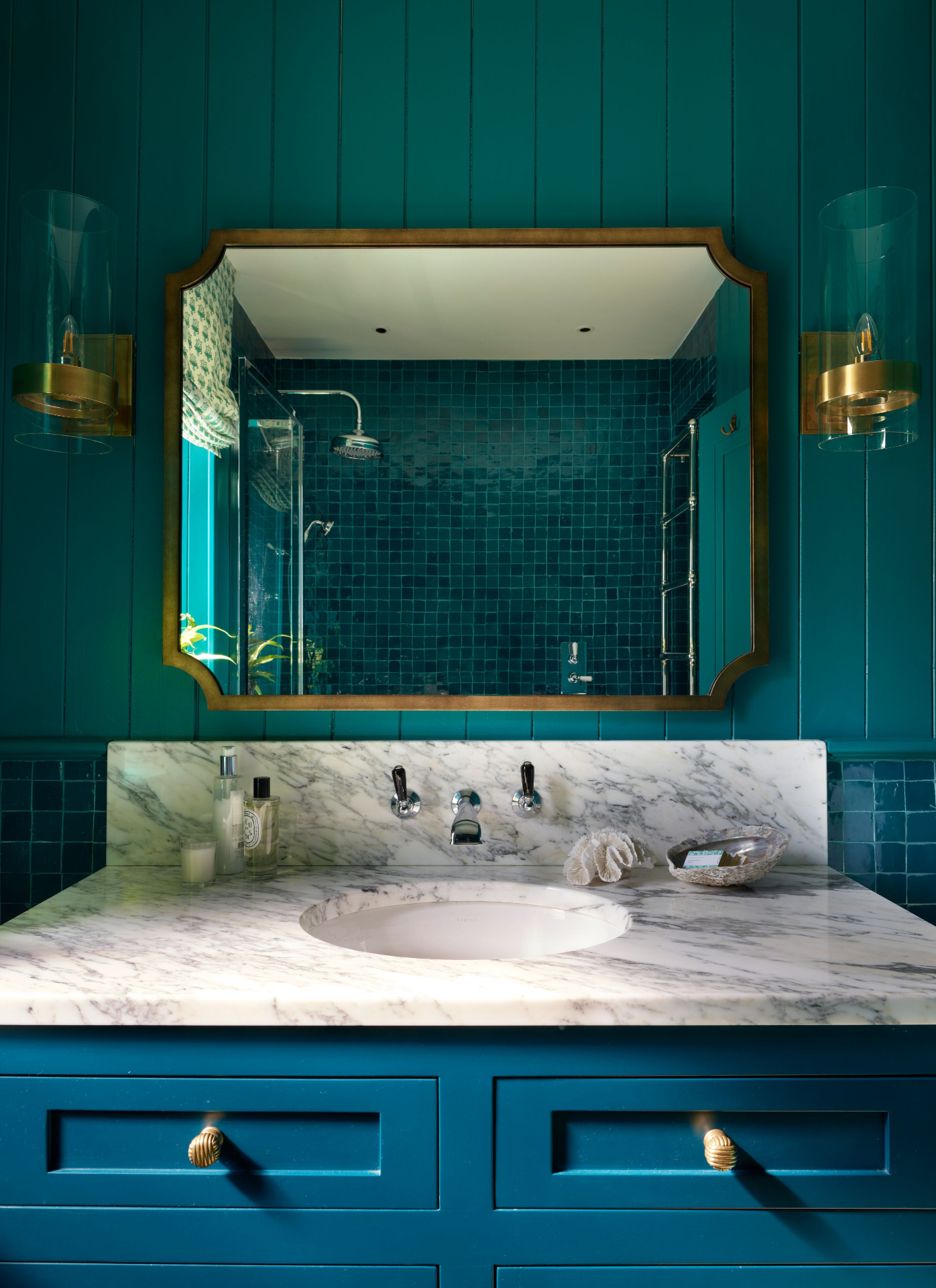 Teal bathroom featuring a blue-green vanity with a white marble countertop, integrated sink, and chrome wall-mounted faucets. Above, a gold-framed mirror reflects tiled shower walls, flanked by two brass-and-glass wall sconces against vertical paneling