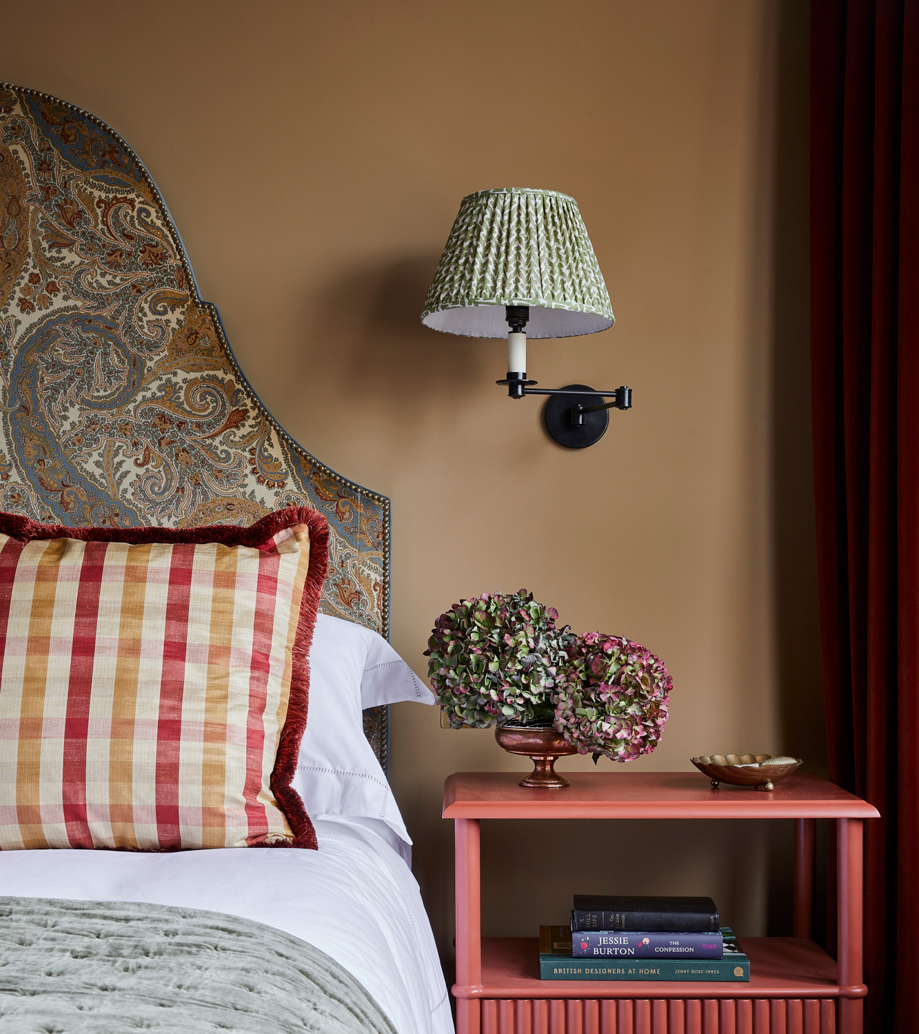 a bedroom with a ochre colour on the wall, a green pleated shade on a wall light, a coral bedside table, a bed with a sage green floral headboard, 