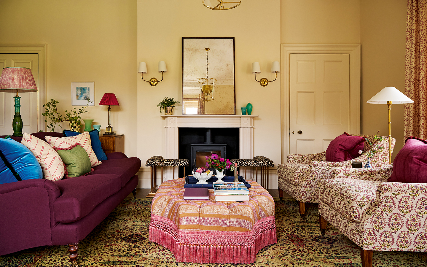 A landscape of a living room with a dark purple sofa on the left, a patterned pink and orange octagonal ottoman in the middle and a pair of patterned purple, yellow and cream armchairs to the right. In the centre is a fireplace topped with a large mirror