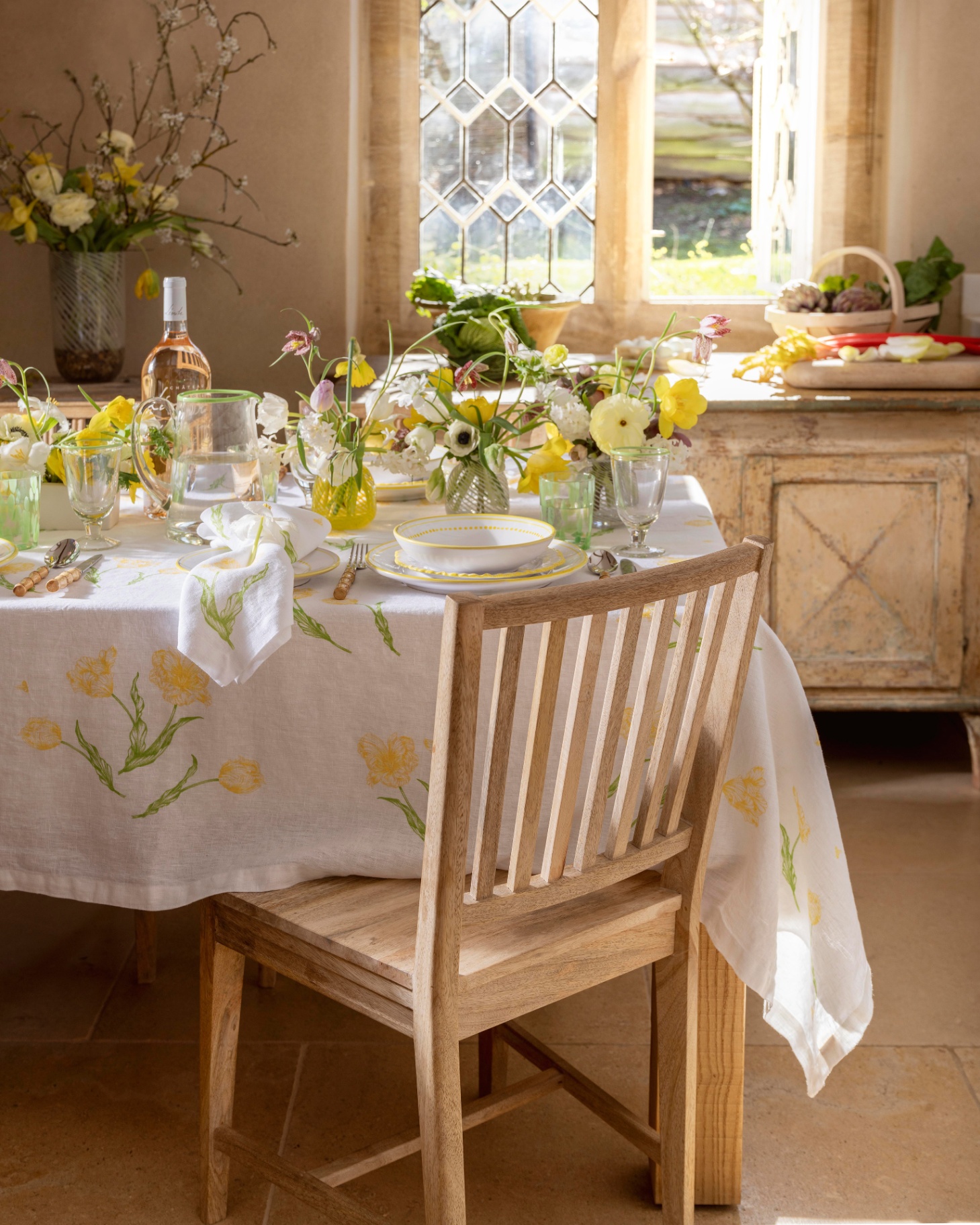 a room with a wooden table and chairs with a white and yellow floral tablecloth and yellow flowers and tableware on top