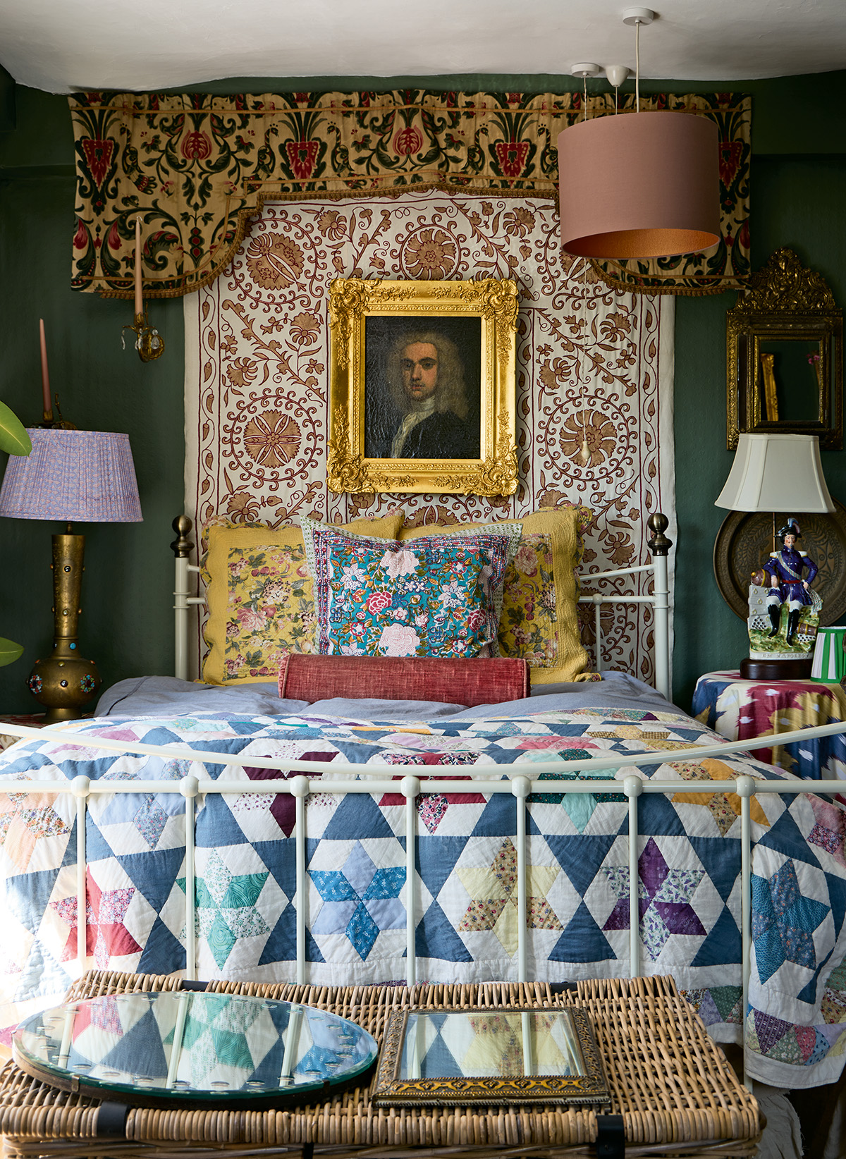 A close up of a bedroom with several clashing patterns. A white wrought iron bed is dressed with floral cushions, bolster cushion and star patchwork quilt. Above hangs a suzani on the wall, layered with a portrait painting. Mismatched lamps sit either side
