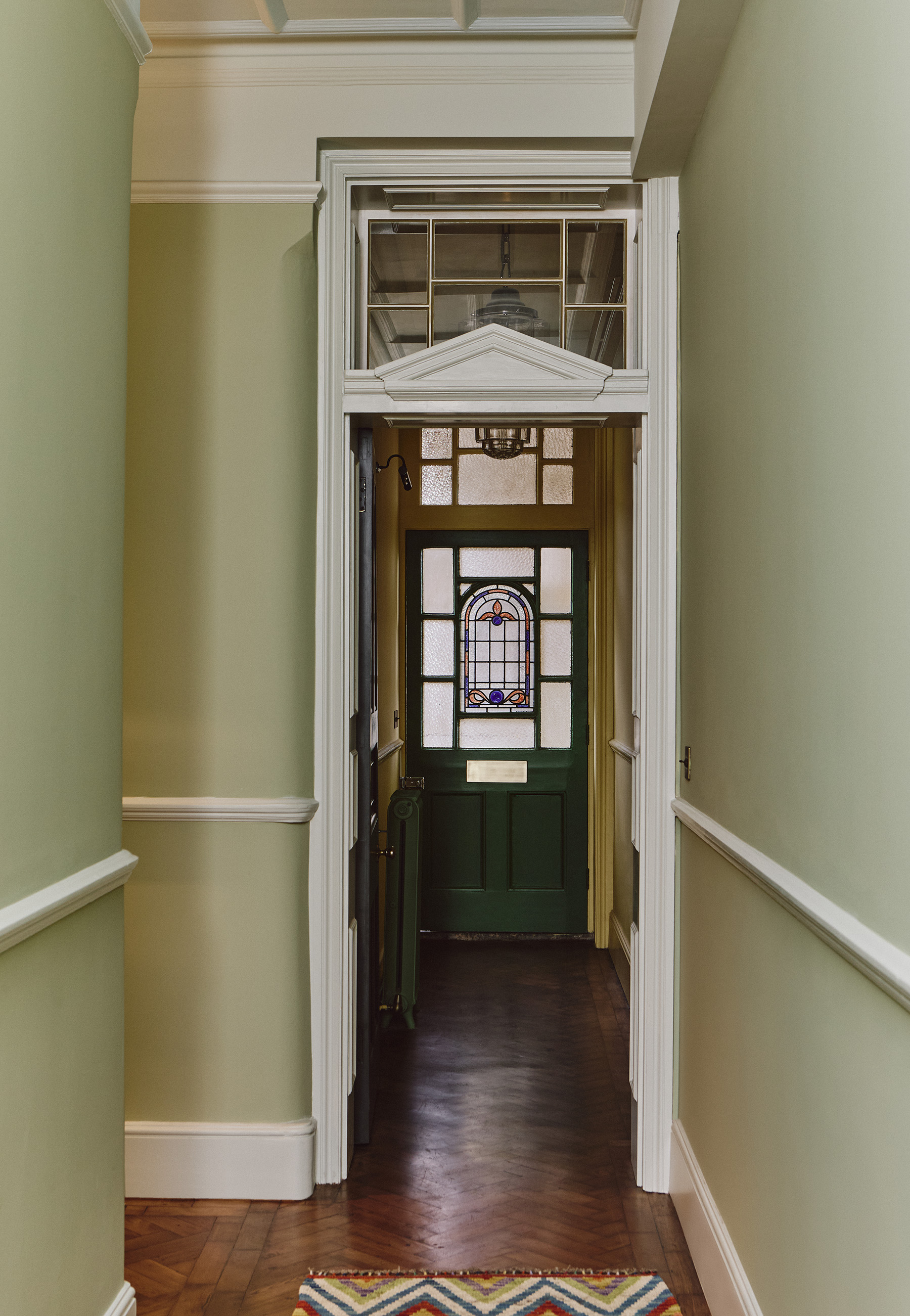 A corridor in an Edwardian flat in London with pale green walls and dark wood floors. There is a doorway surrounded by a white architrave with a pediment detail on top and surrounding a transom window. In the background is the stained glass front door