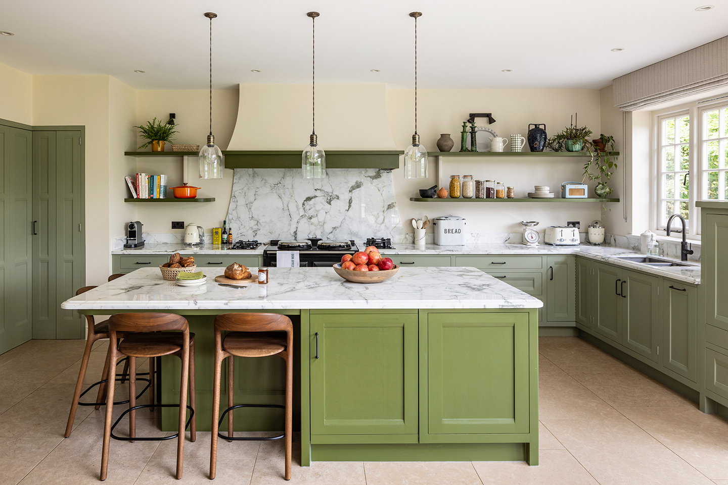 a kitchen with sage green lower cabinet and a brighter green kitchen island with white veined marble worktops. Four wooden bar stools are arranged around one end of the kitchen island and an oven sits in front of a marble backsplash on the back wall