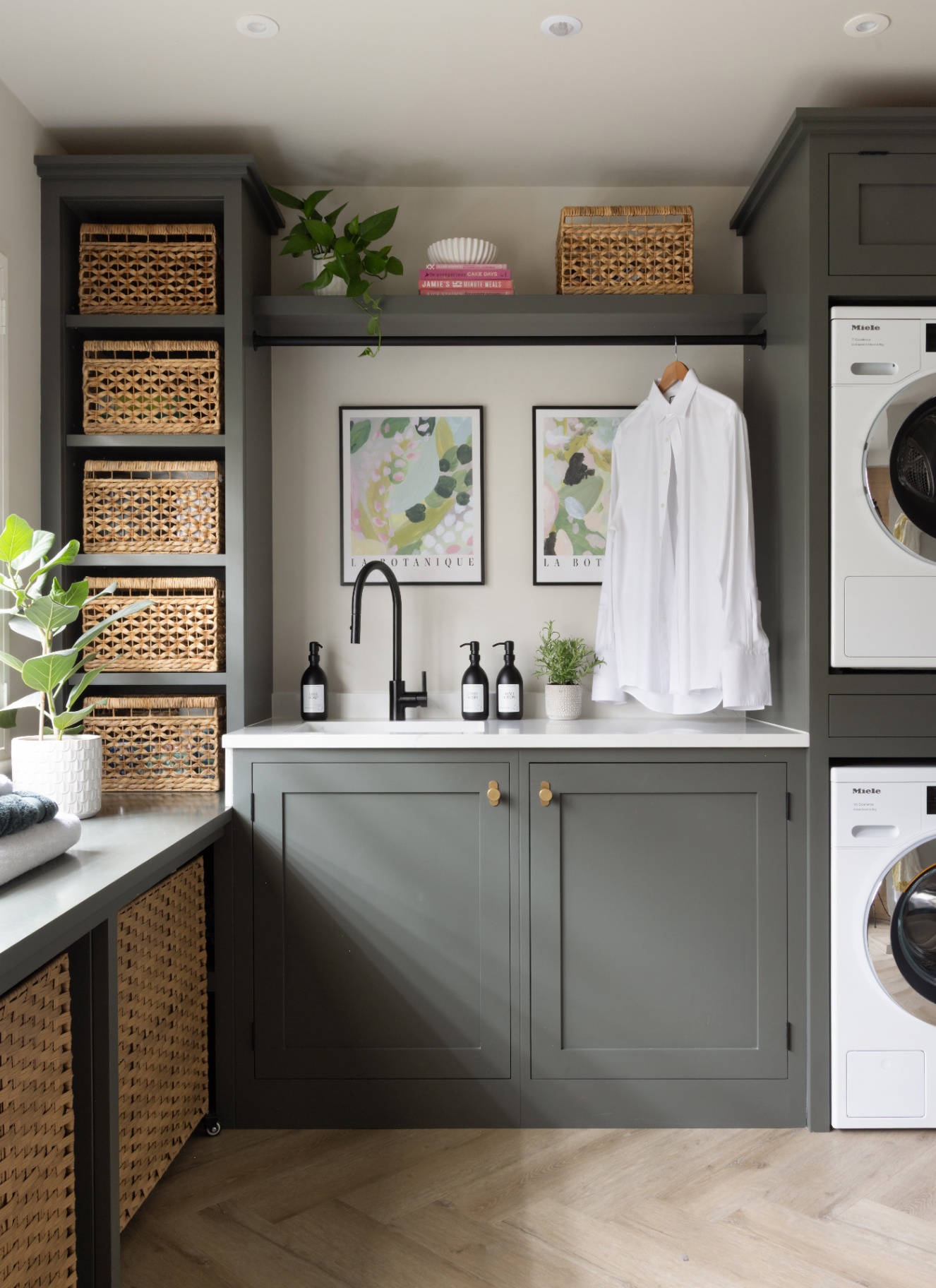 a utility room with green cabinets, shelving alcoves filled with wicker baskets, stacked washer and dryers and a shirt hanging over the sink