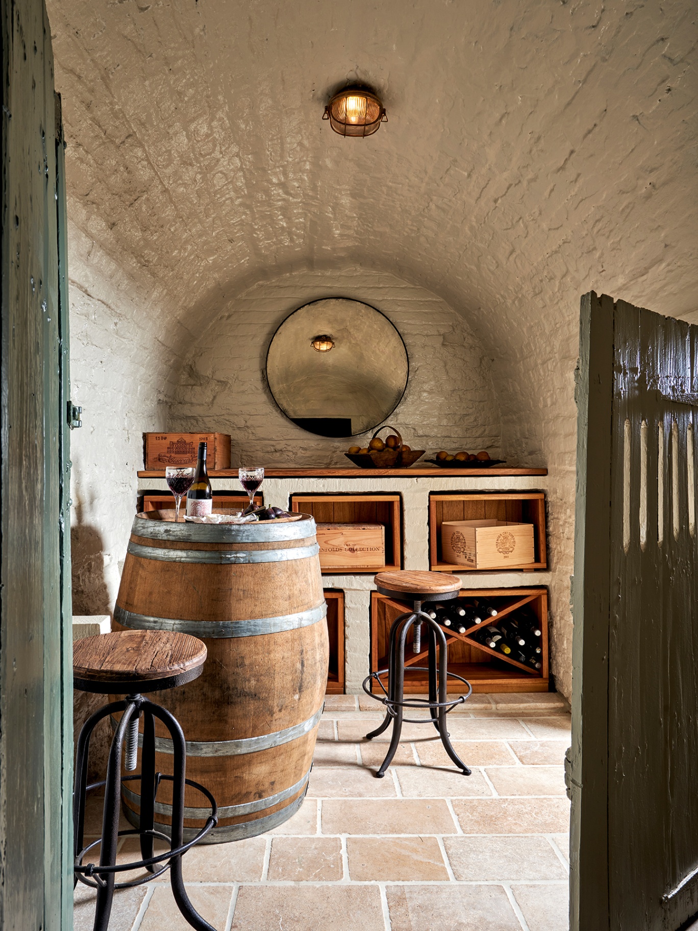 A wine cellar with white painted brick walls and arched ceiling and stone floor. A large barrel, topped with a wine bottle and stem glasses, is paired with industrial-style stools in the foreground