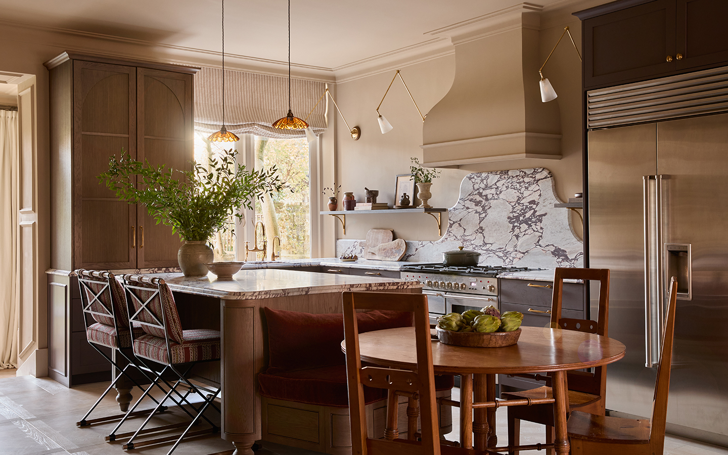a kitchen and dining area with wooden table and chairs in the foreground, a wooden island behind paired with a marble top and two upholstered bar stools, and a surrounding kitchen with dark brown cabinets and dramatic marble worktops
