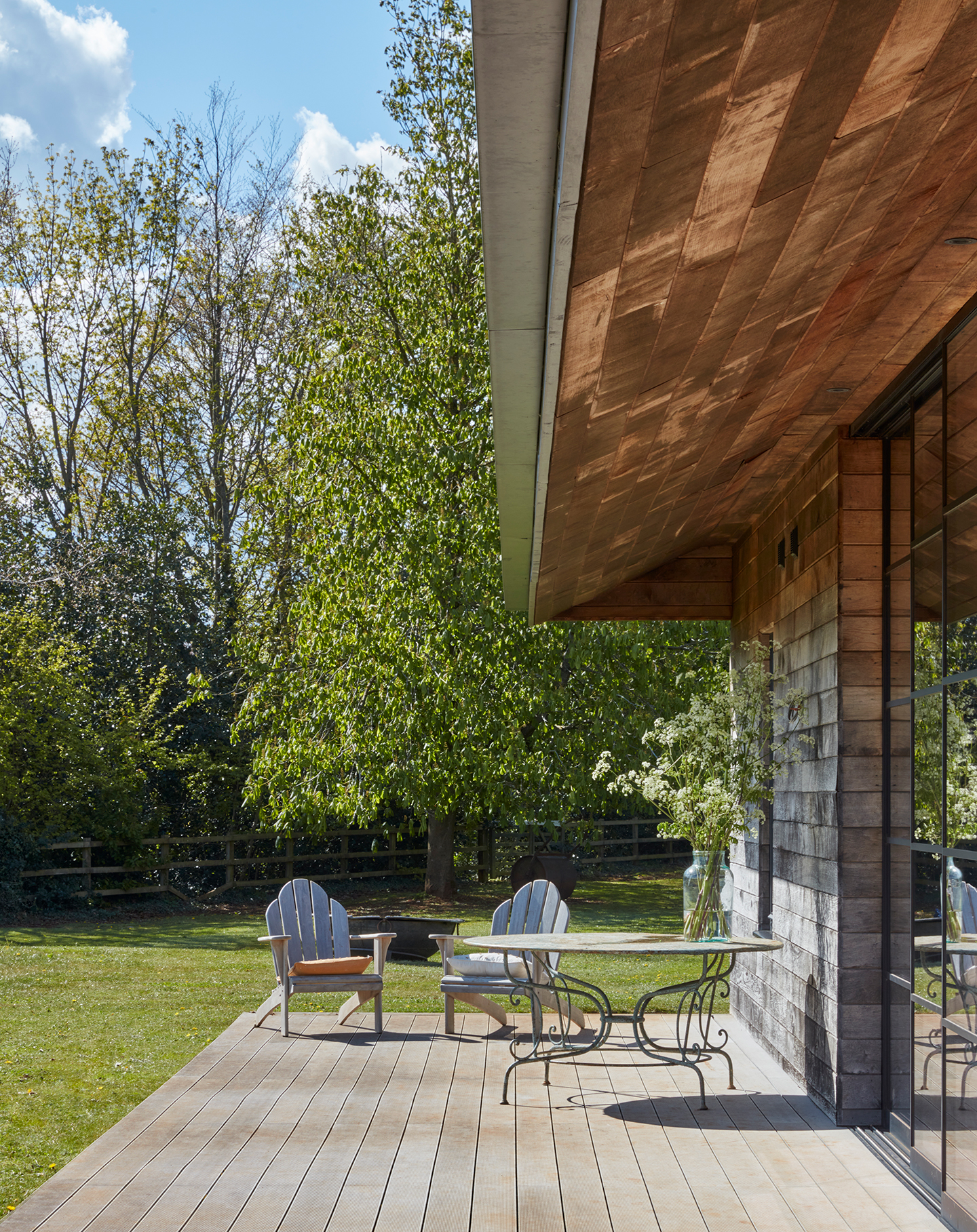 Two wooden chairs and a circular table sit on wooden decking with mature trees and grass in the background. The edge of a house can be seen on the right