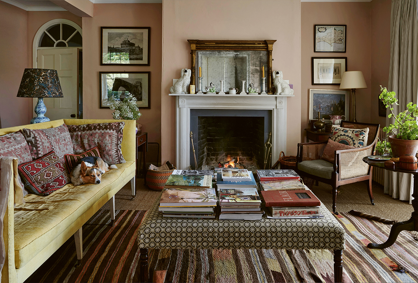 a pale pink living room with a yellow sofa, upholstered ottoman covered in books, armchair and circular table on a striped flatweave rug. On the back wall is a fireplace, mirror and selection of artwork