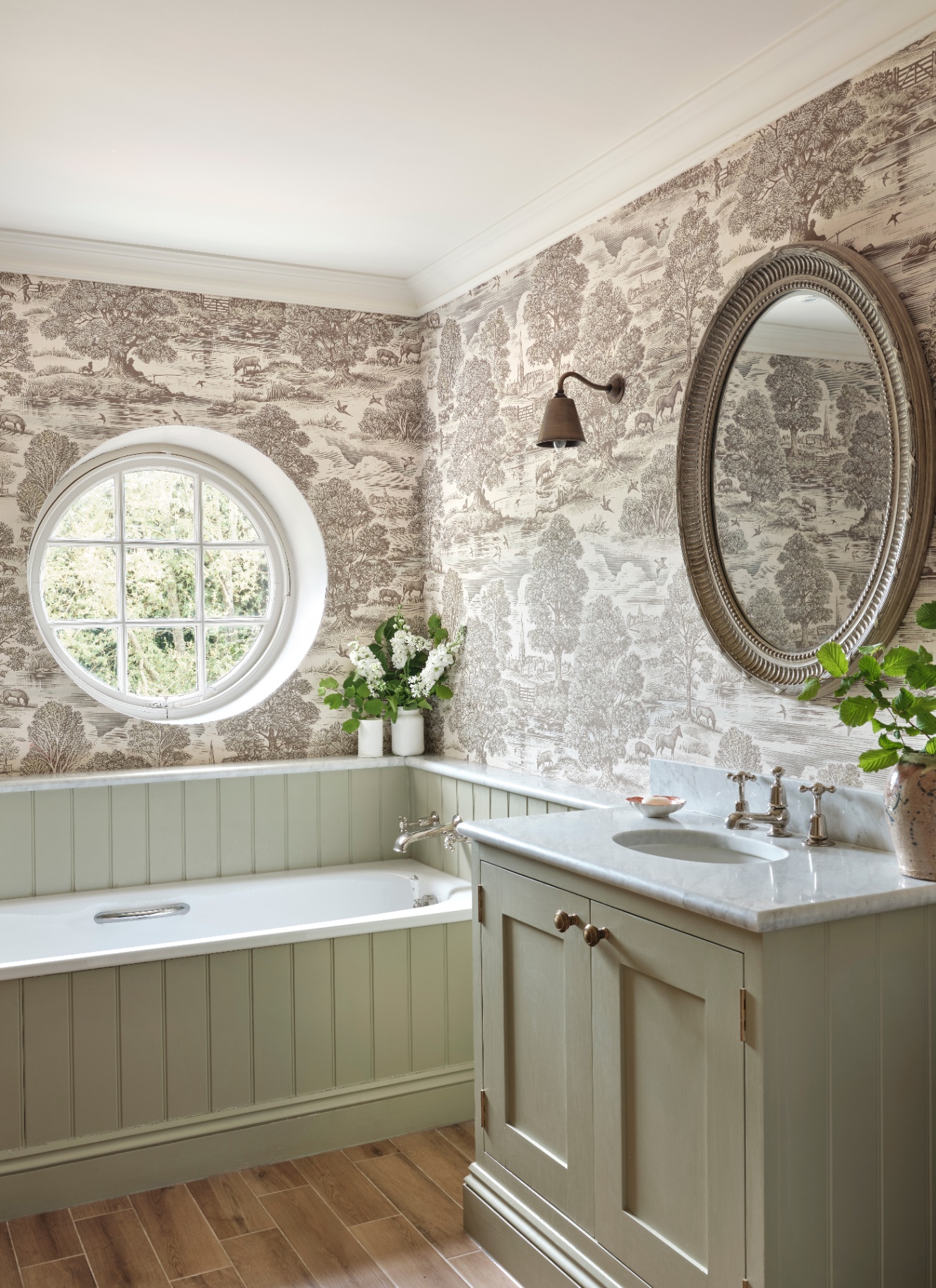 a bathroom with pale sage green panelled bath and vanity unit, grey and white toile de joy wallpaper nad a large circle mirror