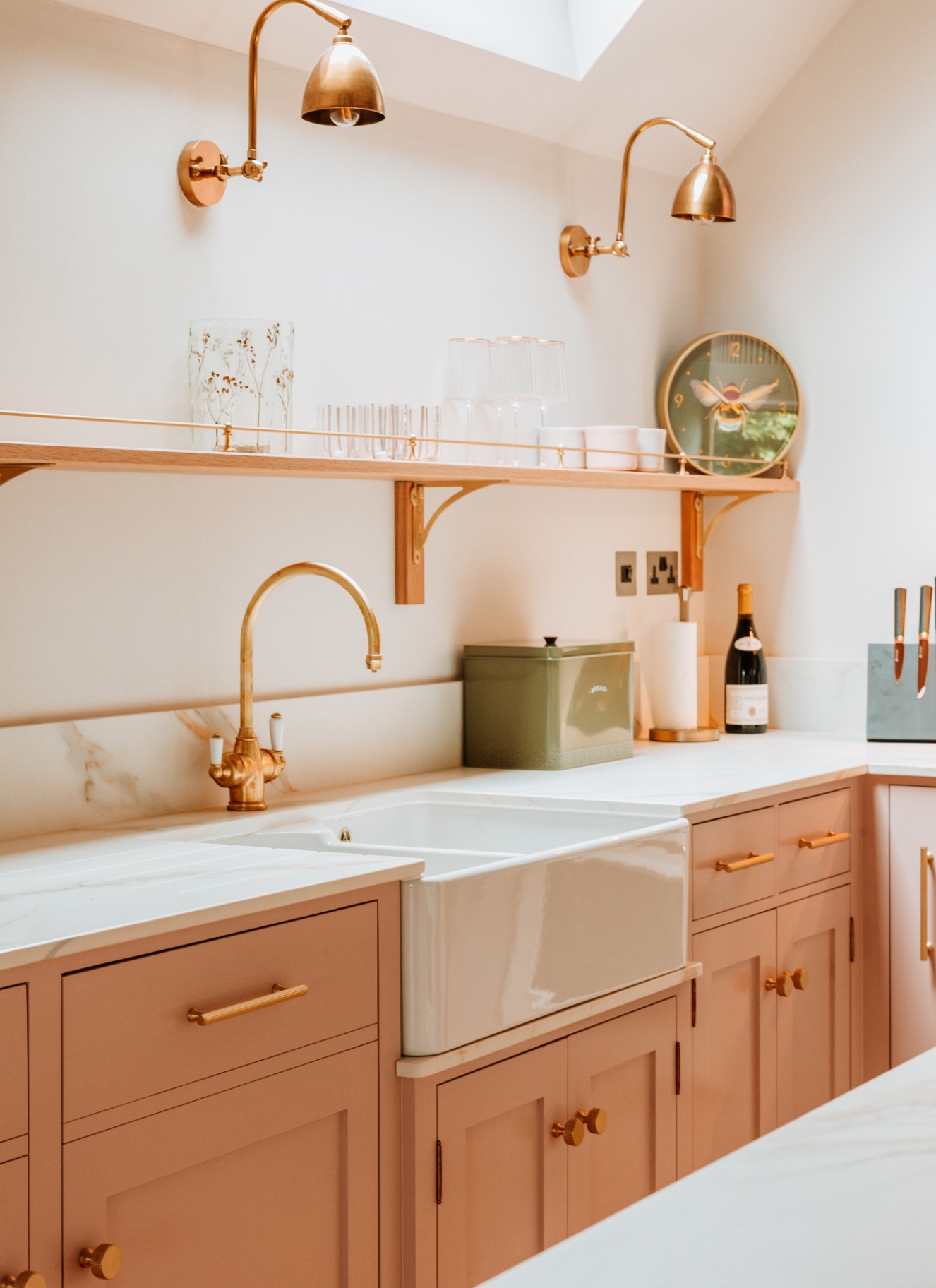 a kitchen sink area with salmon pink cabinets, a belfast sink with a gold tap, a green compost caddy and two task lights above an open shelf