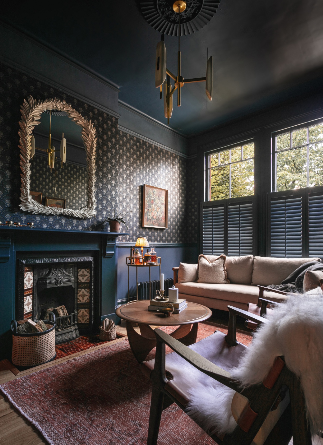 a living room with dark navy walls, a navy fireplace with a large leaf gilded mirror above it, a cream sofa, a small round wooden coffee table and a modern light fitting