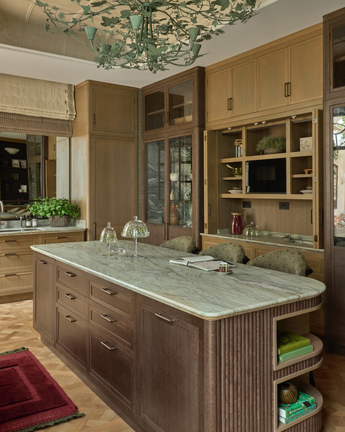 a kitchen with a dark wooden kitchen island with a grey marble top, a large green ornate iron light fixture in the shape of leaves and wooden cabinetry