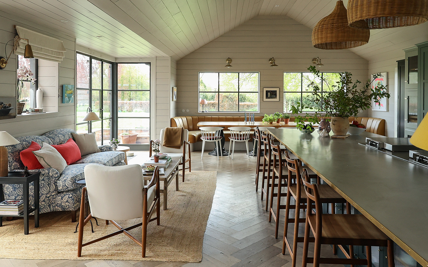 A large open-plan kitchen-dining-living room with a large island on the right with rattan pendants hanging above, a dining table with chairs and banquette seating at the back, and a seating area consisting of a sofa, coffee table and chairs to the left