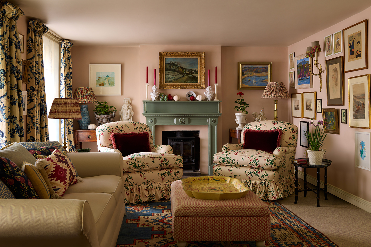 A pretty pink living room with a green fireplace in the centre of the back wall. Two patterned frilled armchairs sit either side, and there is a central ottoman and sofa to the left. The walls are covered with art