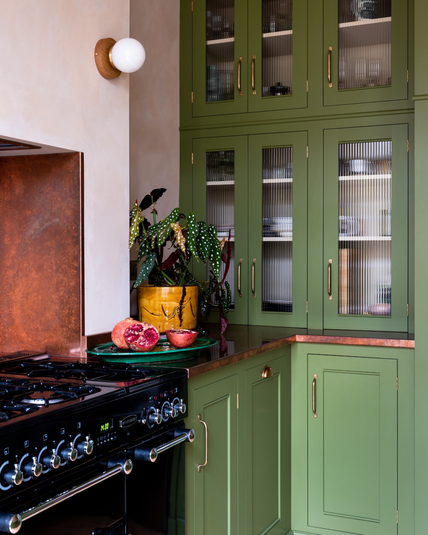 a kitchen with green cabinetry, a black cooker, a copper splashback, copper worktops, a yellow plant pot with a green plant inside and a spherical wall light