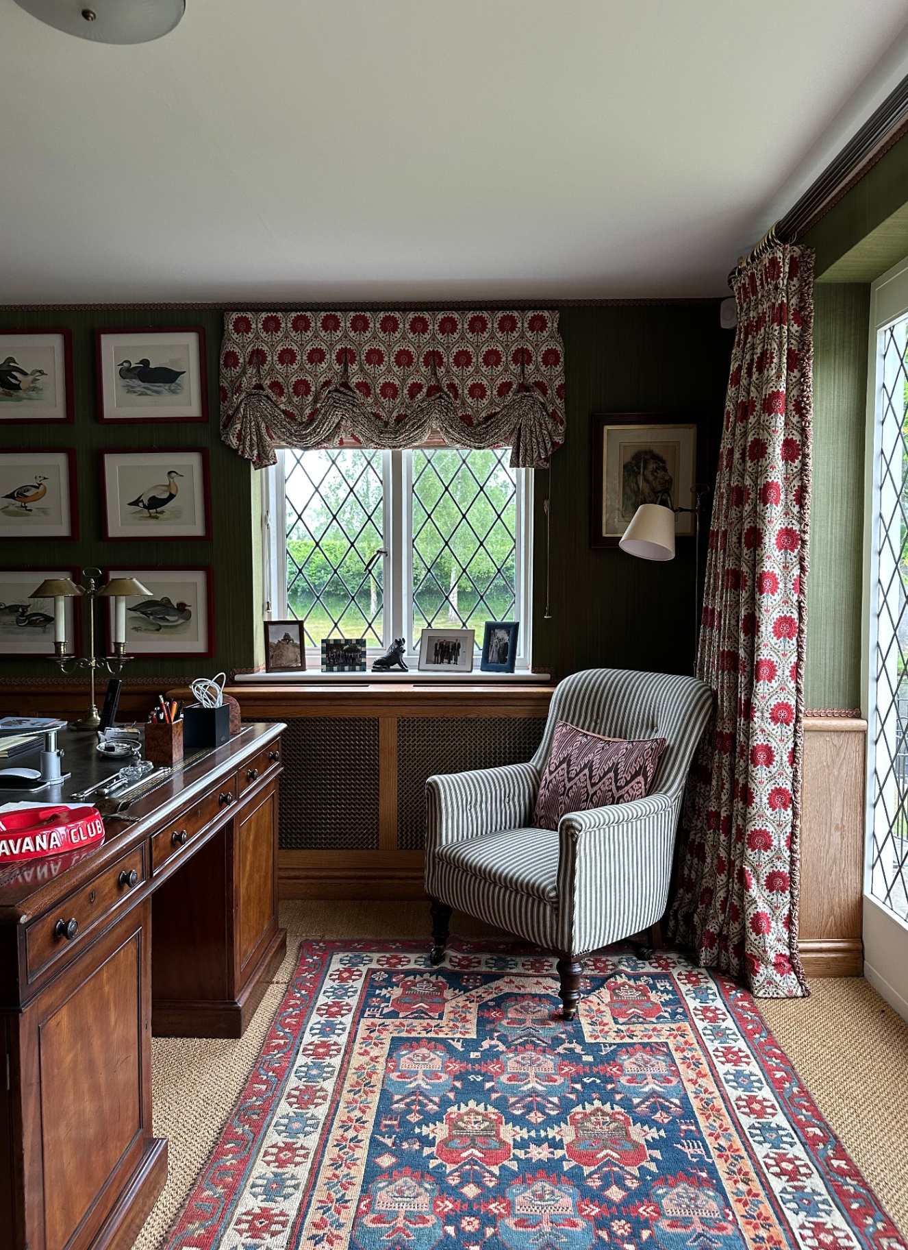 Traditional study with dark green walls, leaded casement windows framed by floral full-length curtains and Roman blinds, a dark wood desk, and a striped wingback armchair