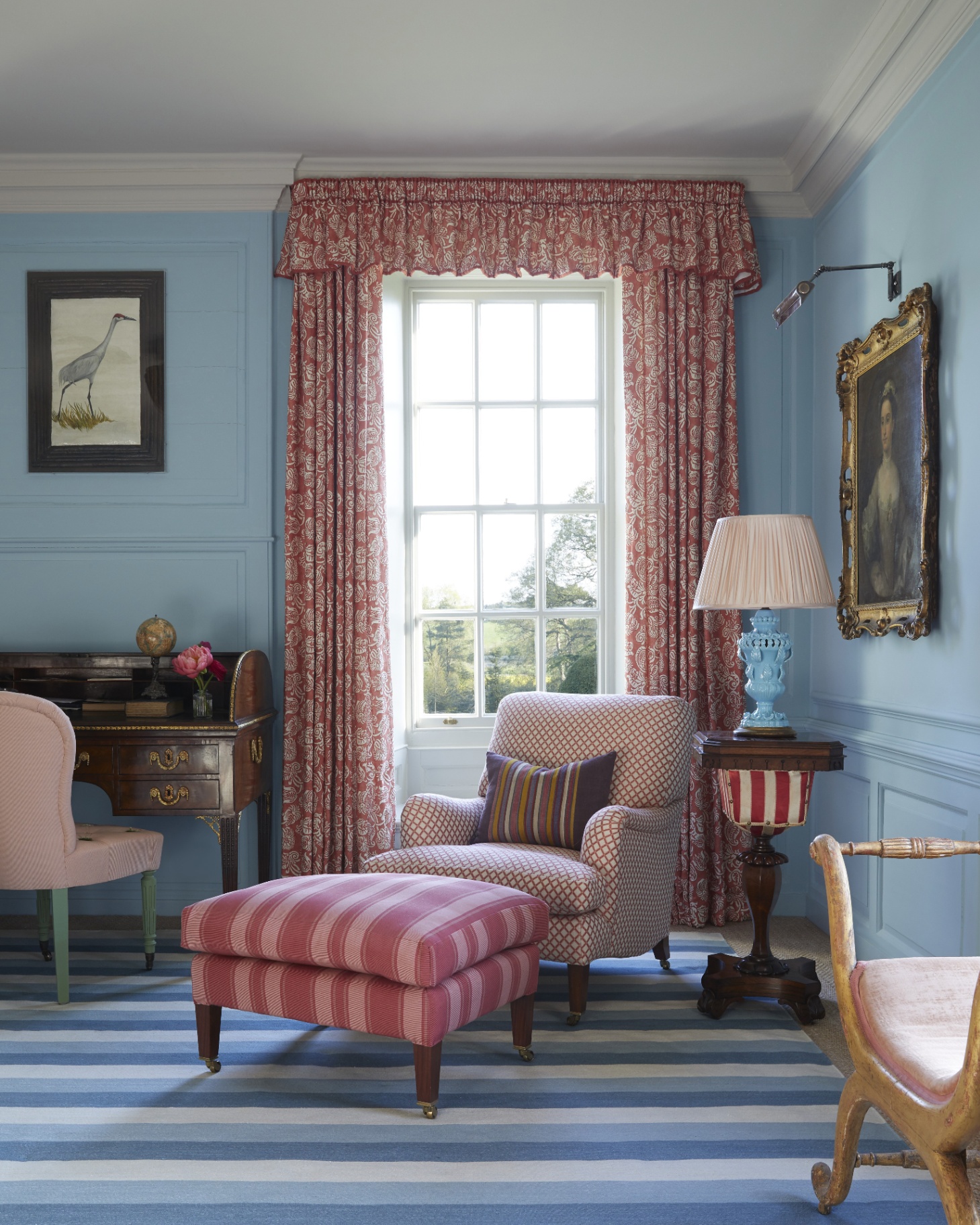 a living room corner with pale blue painted walls, a blue stripe rug, a windown with orange and white floral curtains, an armchair with a pink stripe footstool and an antique desk