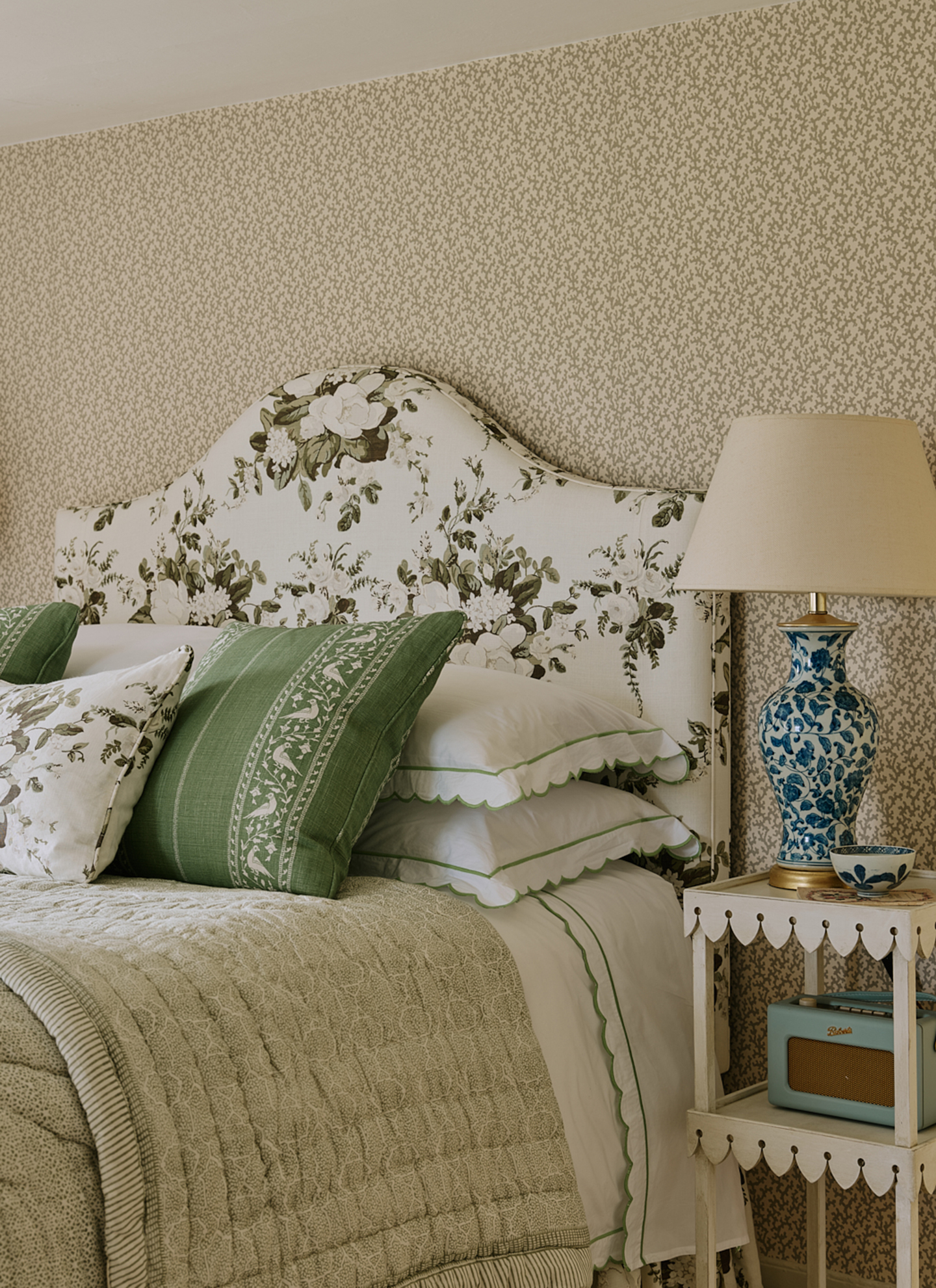 a bedroom with white and green leaf print wallpaper, a bed with a floral headboard and a green bedspread and a small scalloped beside table with a blue and white floral lamp
