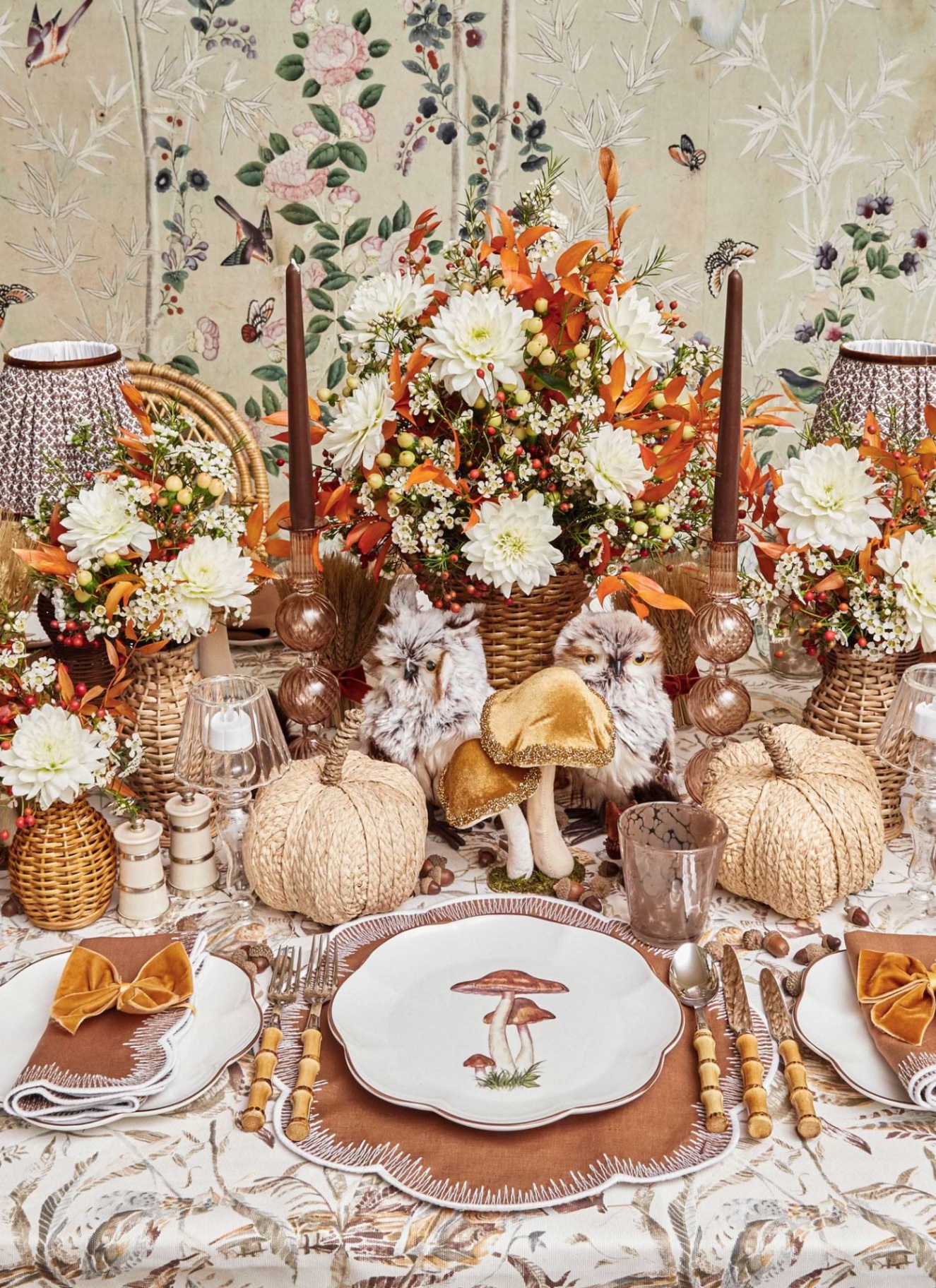 an autumn tablescape with rattan pumpkins, a vase of white and orange flowers, velvet mushrooms, brown tapered candles, a plate with a mushroom illustration, bamboo cutlery and a brown scalloped placemat