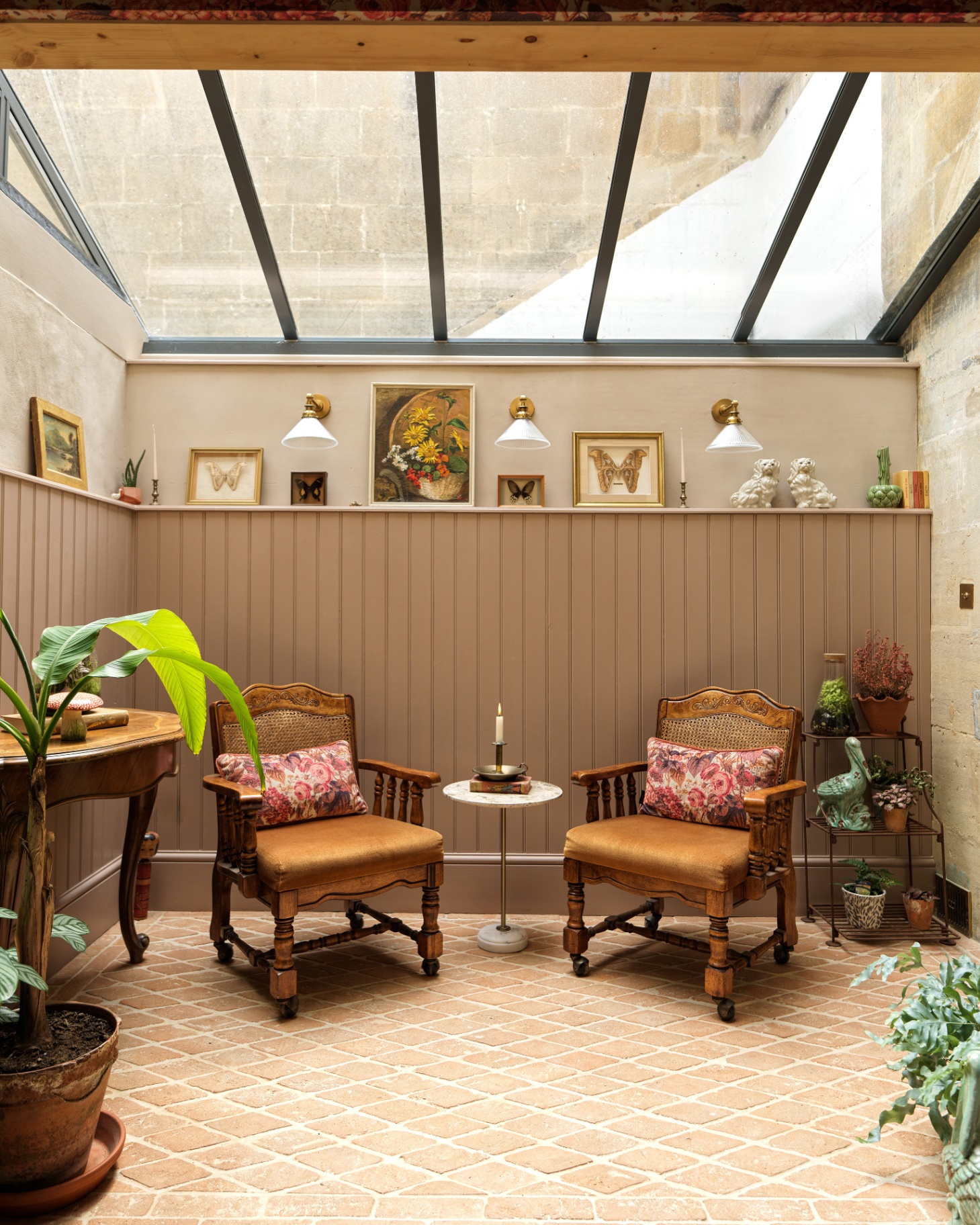 a conservatory room with beige paneled walls with ledge shelves filled with decorative items, two brown antique armchairs with cushions and teracotta tiled floors