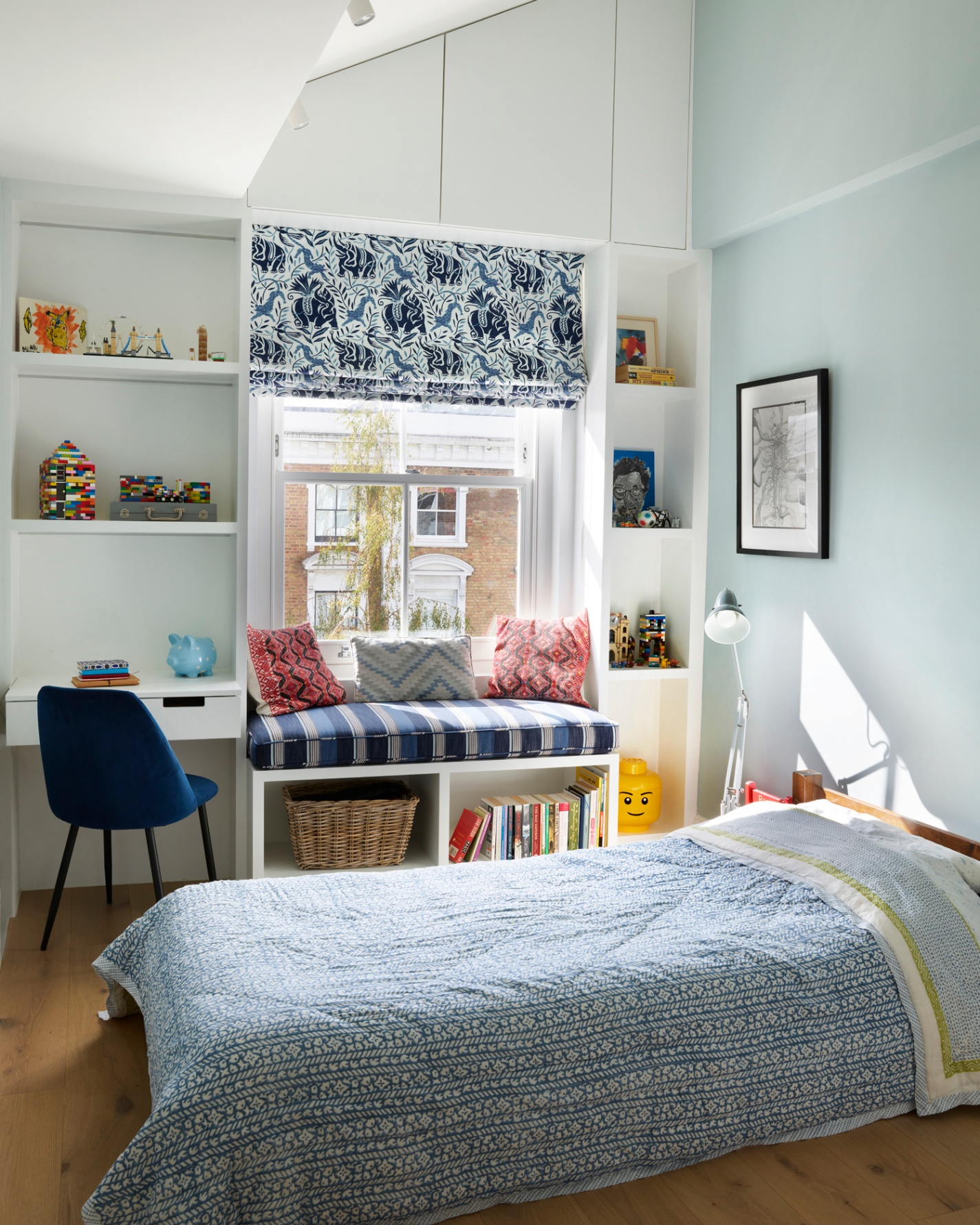 a children's bedroom with a window seat, shelves filled with toys, a small built in desk and a low single bed with a white and blue patterned bedspread