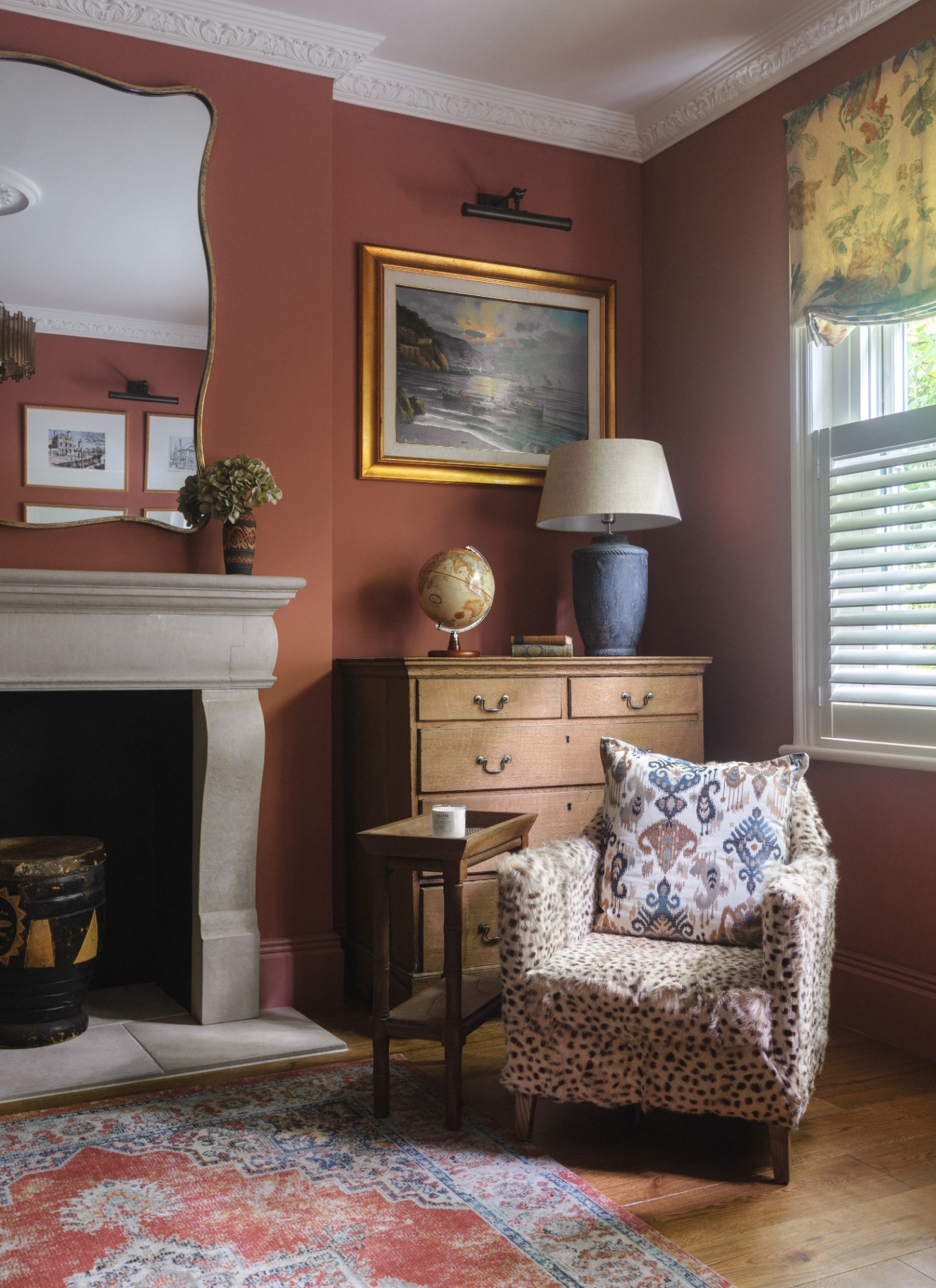 a living room corner with terracotta walls, a fluffy animal print armchair, a wooden chest of drawers behind the chair and a large stone fireplace with a wavy mirror above it