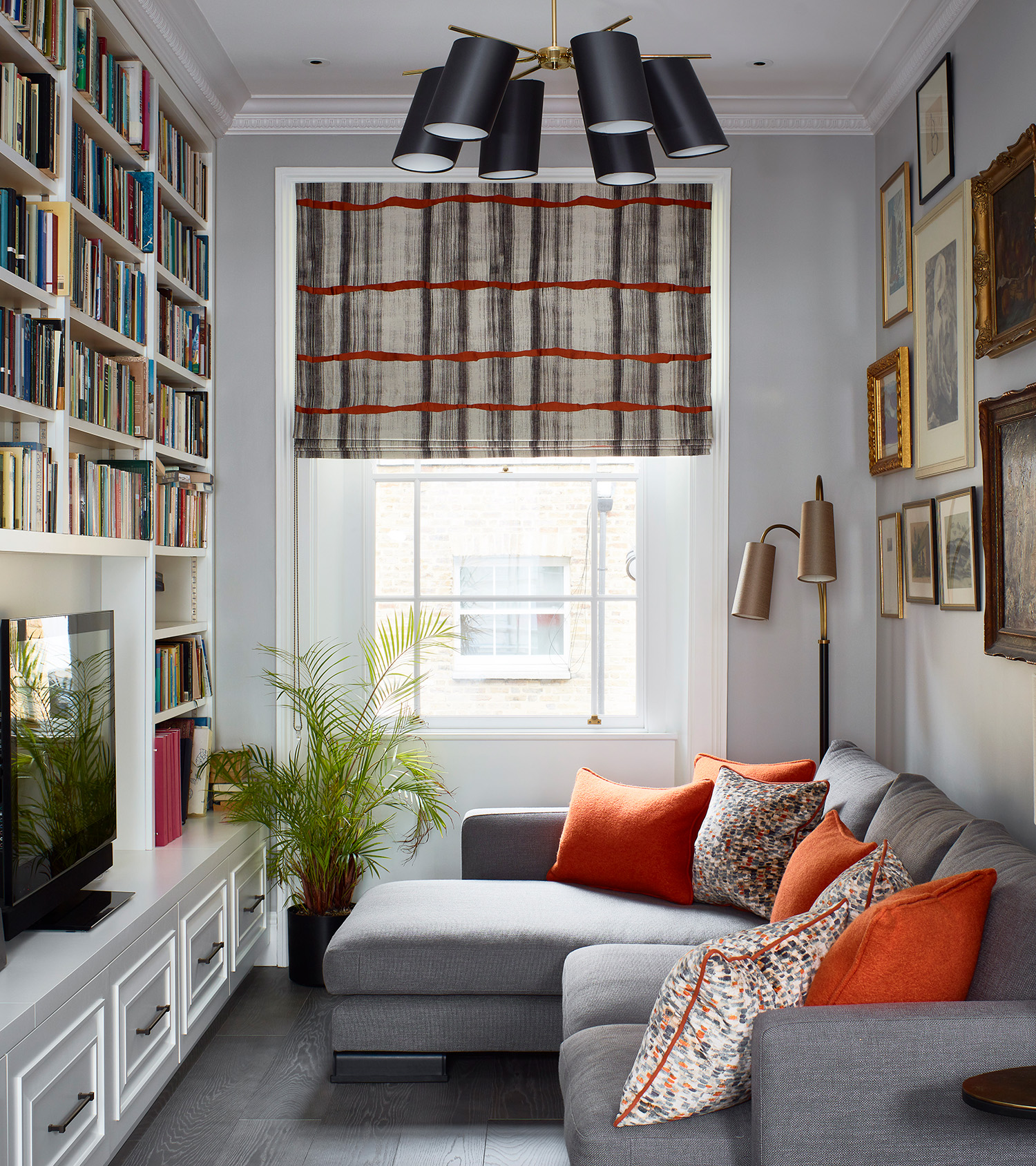 a small living room with pale grey walls, a window with a grey and orange patterned blind, a grey chaise sofa with grey and orange cushions and a gallery wall above and a floor to ceiling bookcase opposite the sofa with a tv in the middle