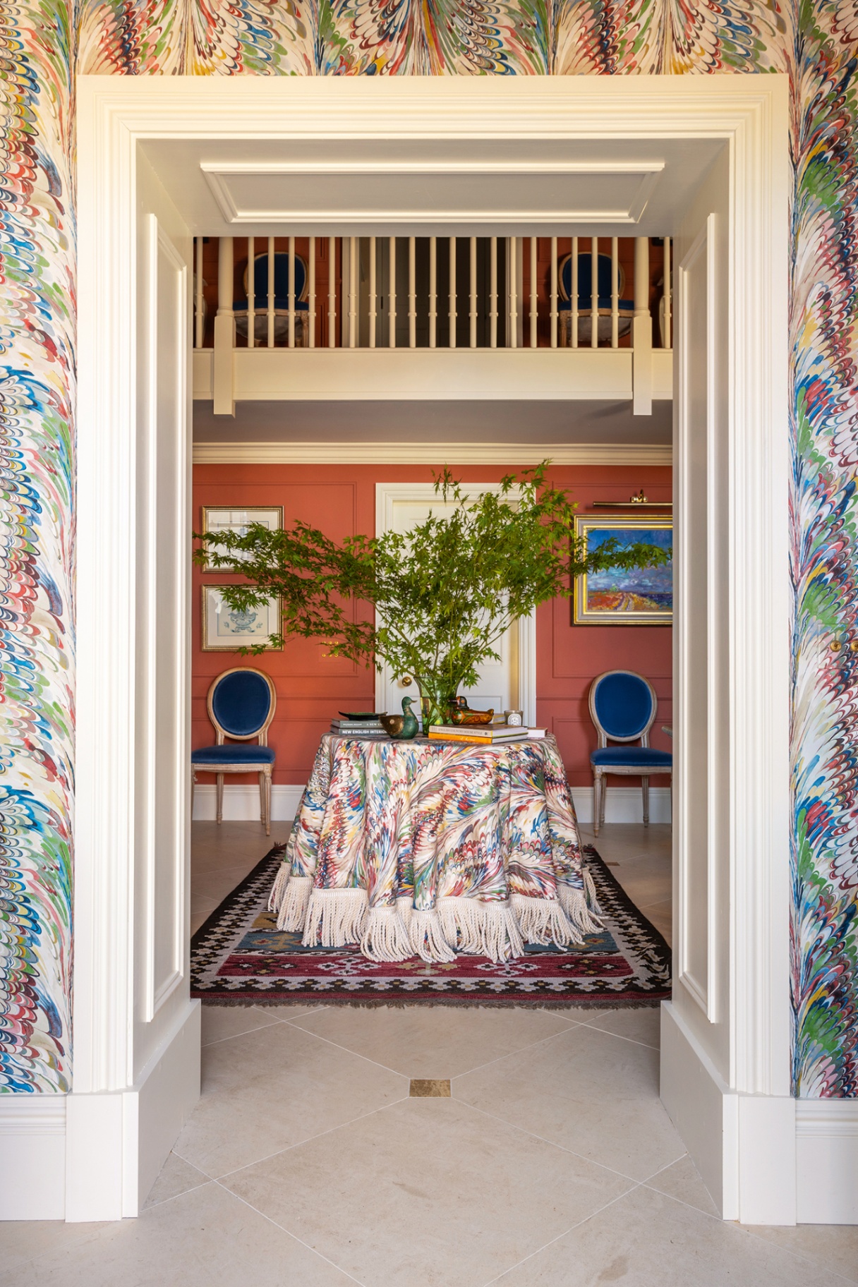 A view through to a grand country house hall where a circular table, topped with a marbled fabric tablecloth with tassels, sits on an antique rug. Matching wallpaper can be seen in the foreground and the back walls is painted bright orange