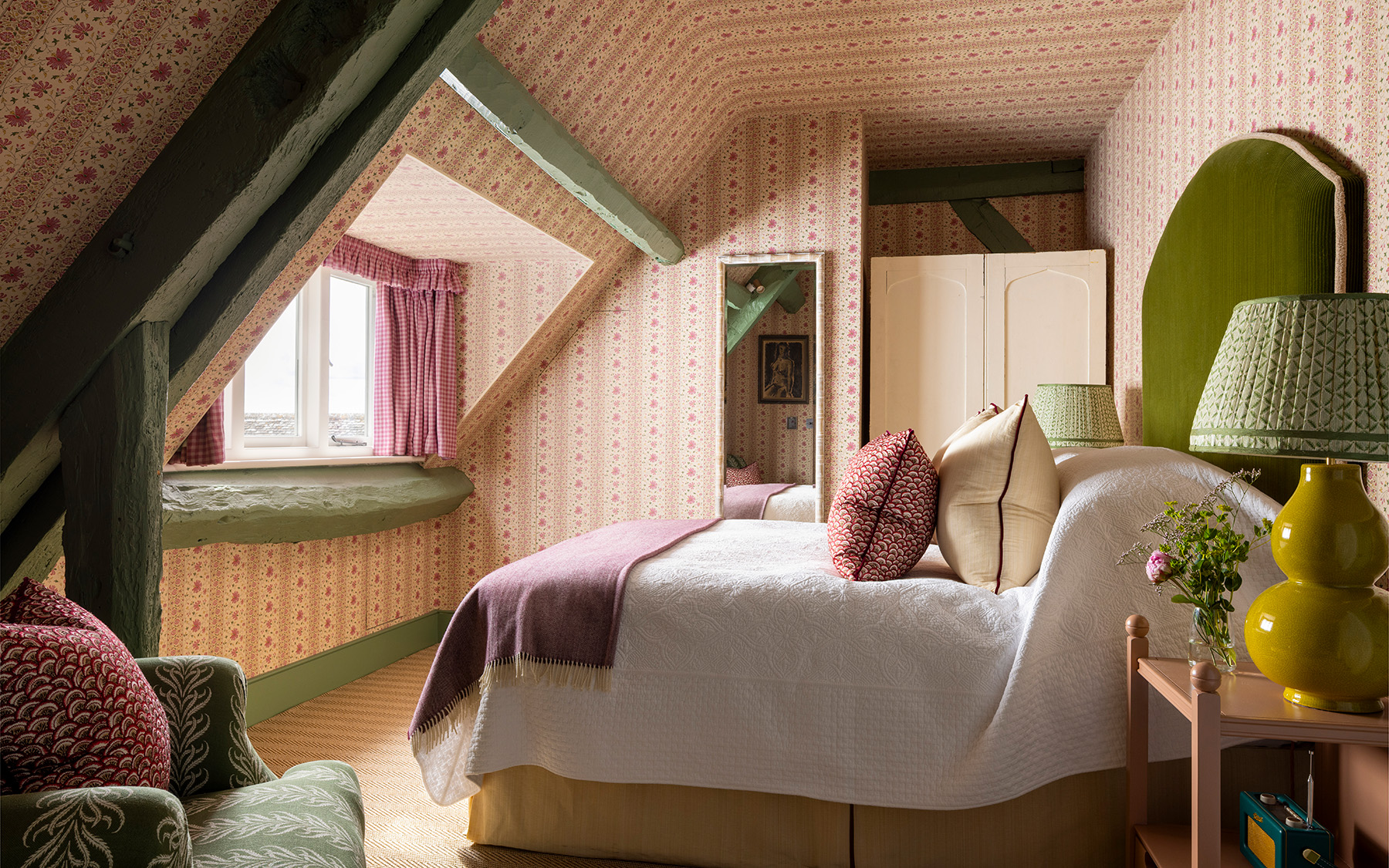 a loft bedroom with green ceiling beams, pink ditsy floral wallpaper on the walls and ceiling and a double bed with an olive green velvet headboard