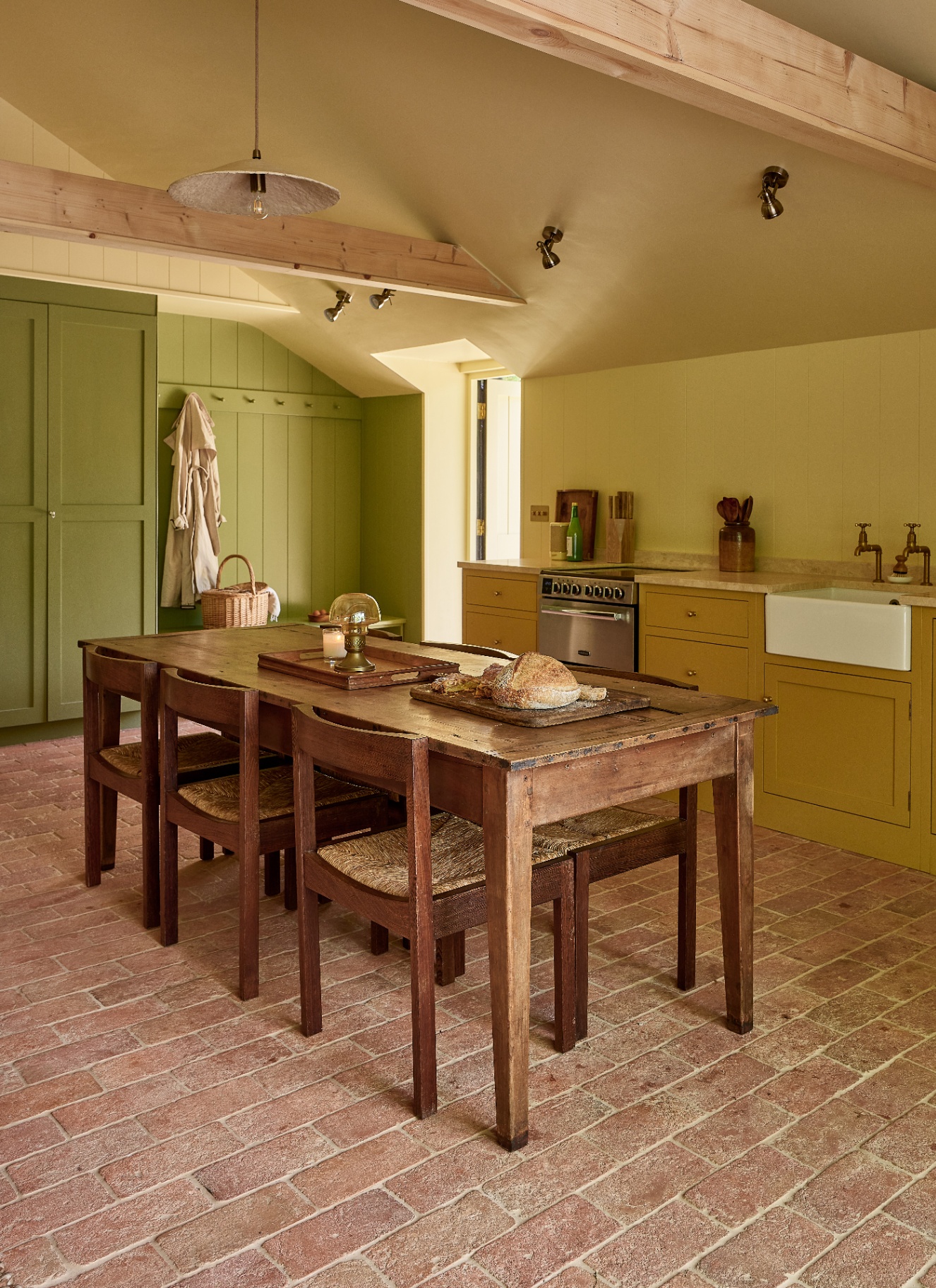 a country kitchen with bricks floors, a large wooden dining table and chairs, mustard yellow cabinetry and green cabinetry at the back