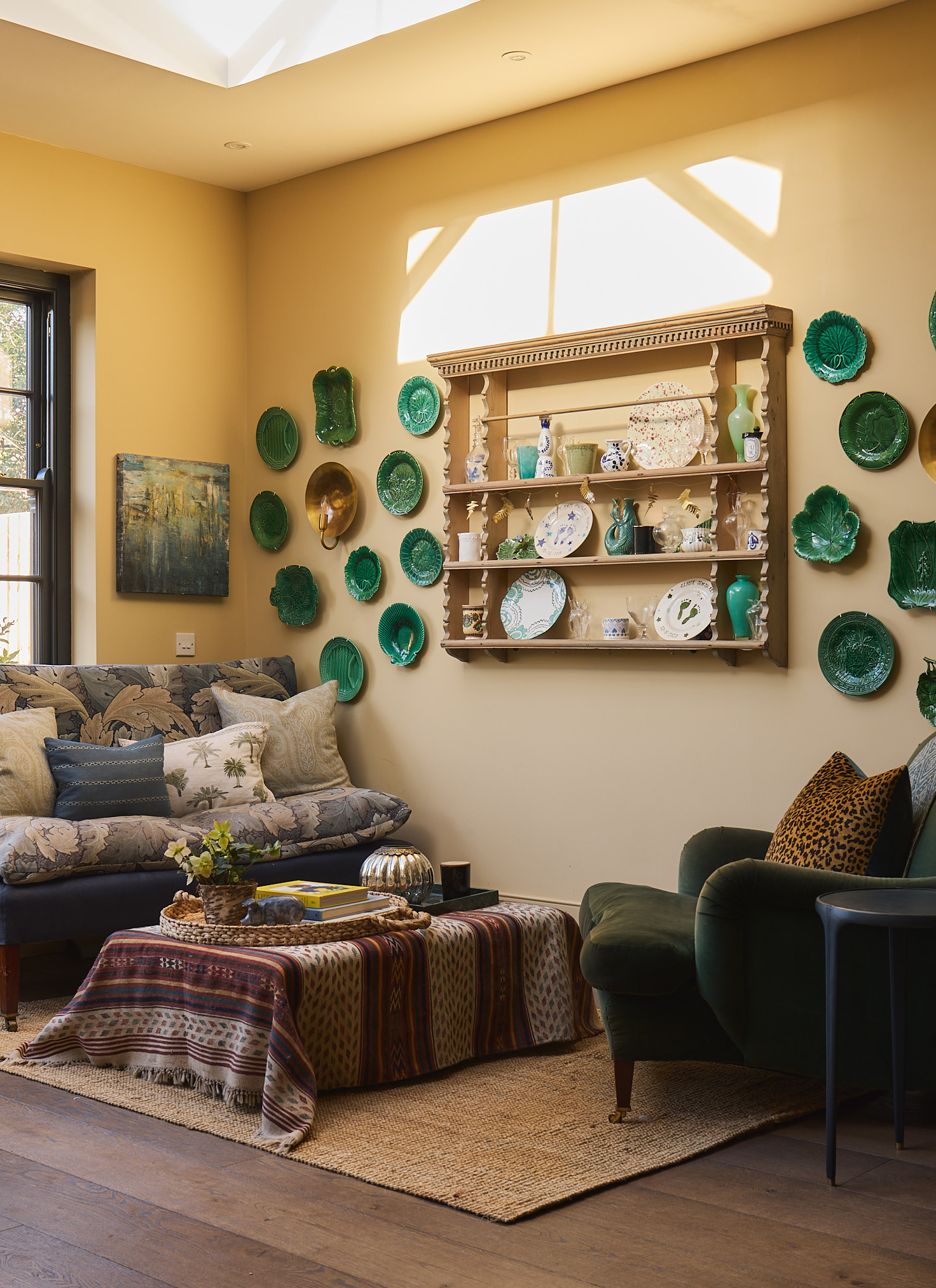a sitting room with pale yellow walls, green plates hung on the wall, an open shelving unit, a jute rig and a blue and a green armchair