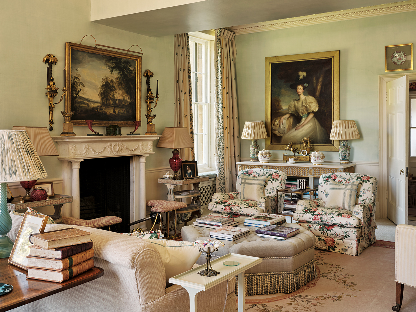 an angled shot of a classic country living room with a cream sofa and patterned floral armchairs surrounding an ottoman stacked with books. The blue-painted walls are decorated with classical paintings and there is a fireplace to the left
