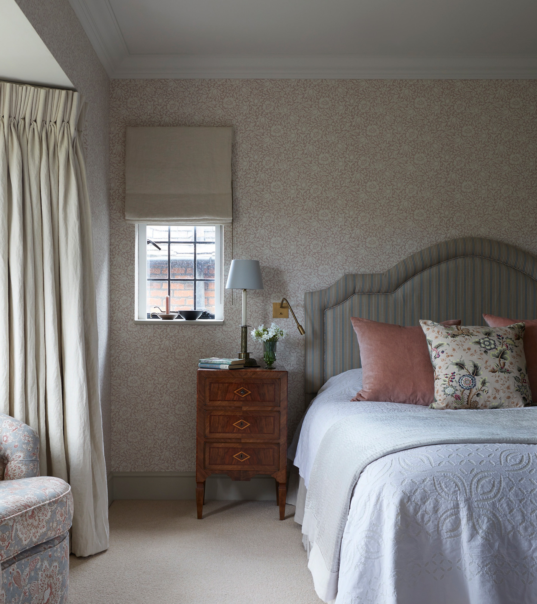 A bedroom with cream curtains, neutral floral wallpaper, a dark wood bedside table and a double bed with a striped headboard, white bed linen and pink and floral cushions