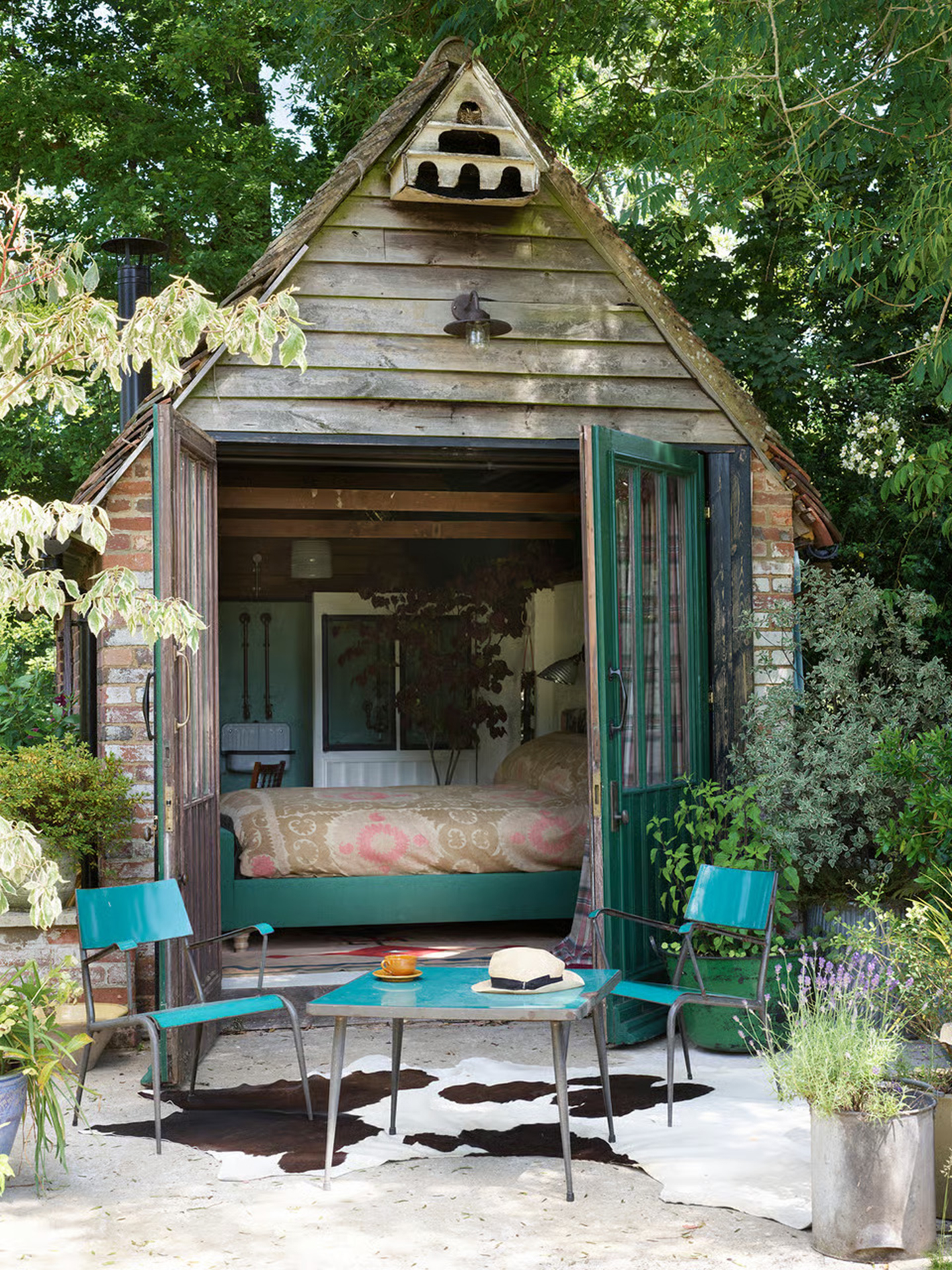 an outhouse with a turqoise blue bistro set outside with a cowhide rug and a teal bed insde with a beige and pink patterned bed cover