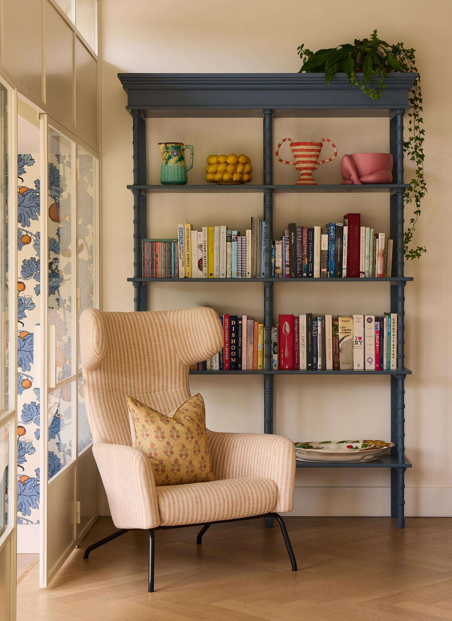 A high-backed pink striped wing chair set beside a tall, architectural blue freestanding bookcase styled with books and ceramic vases.