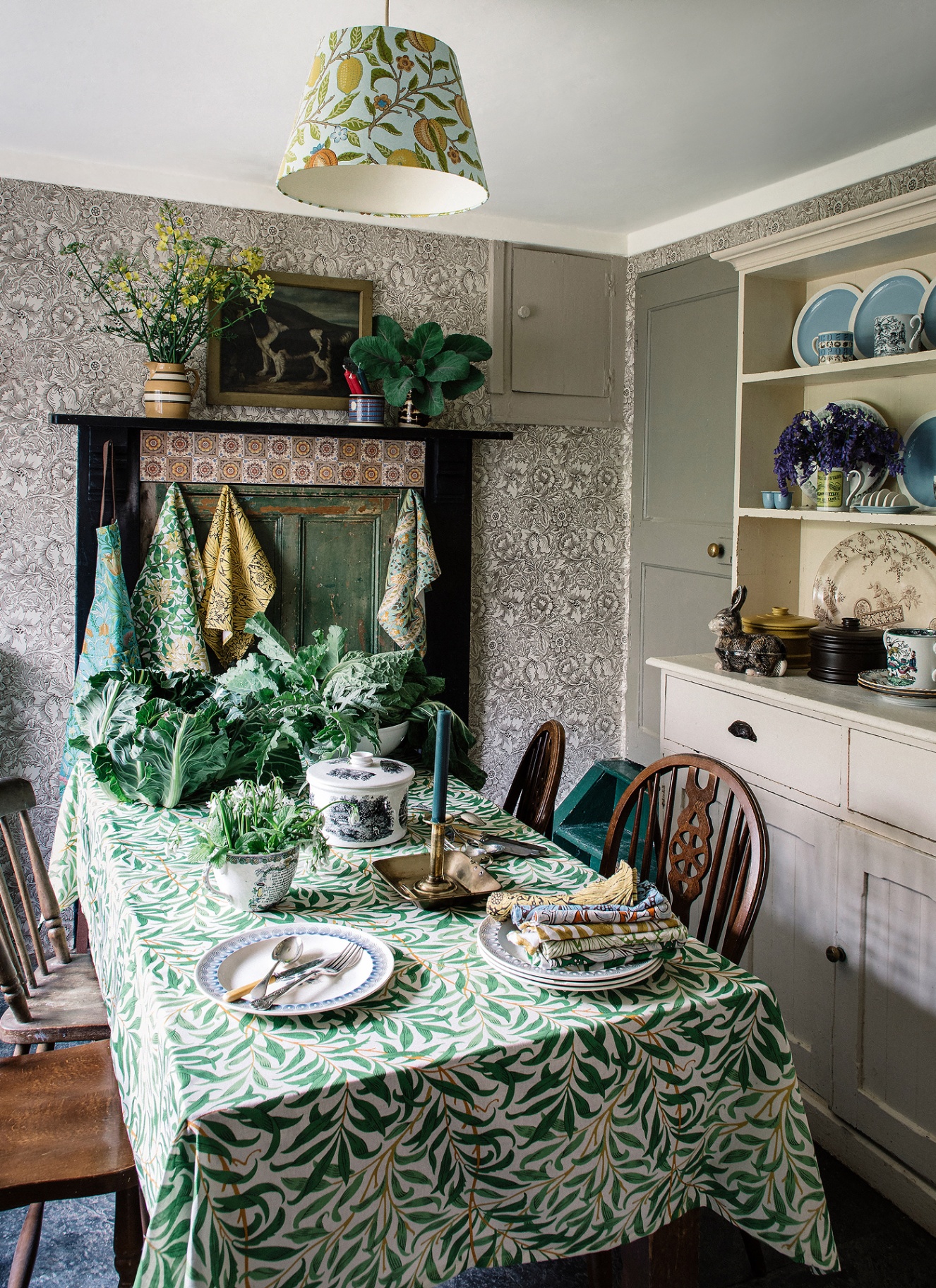 Small cottage dining nook with botanical floral wallpaper, a green leaf patterned tablecloth, vintage wooden chairs, and a cream dresser display