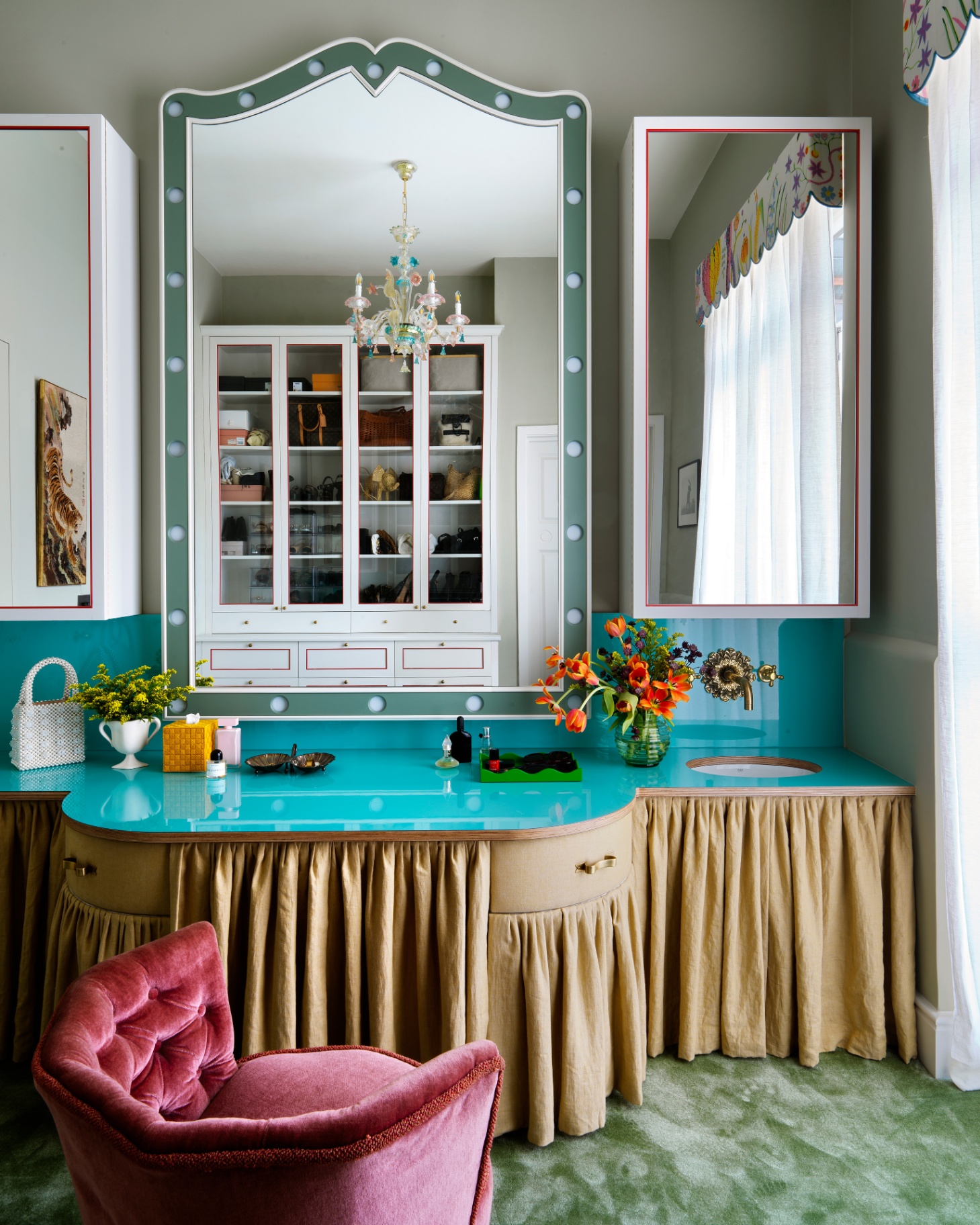 a dressing table with a large mirror with lights above it, a yellow skirt beneath the table, teal worktop and a dark pink velvet tub chair