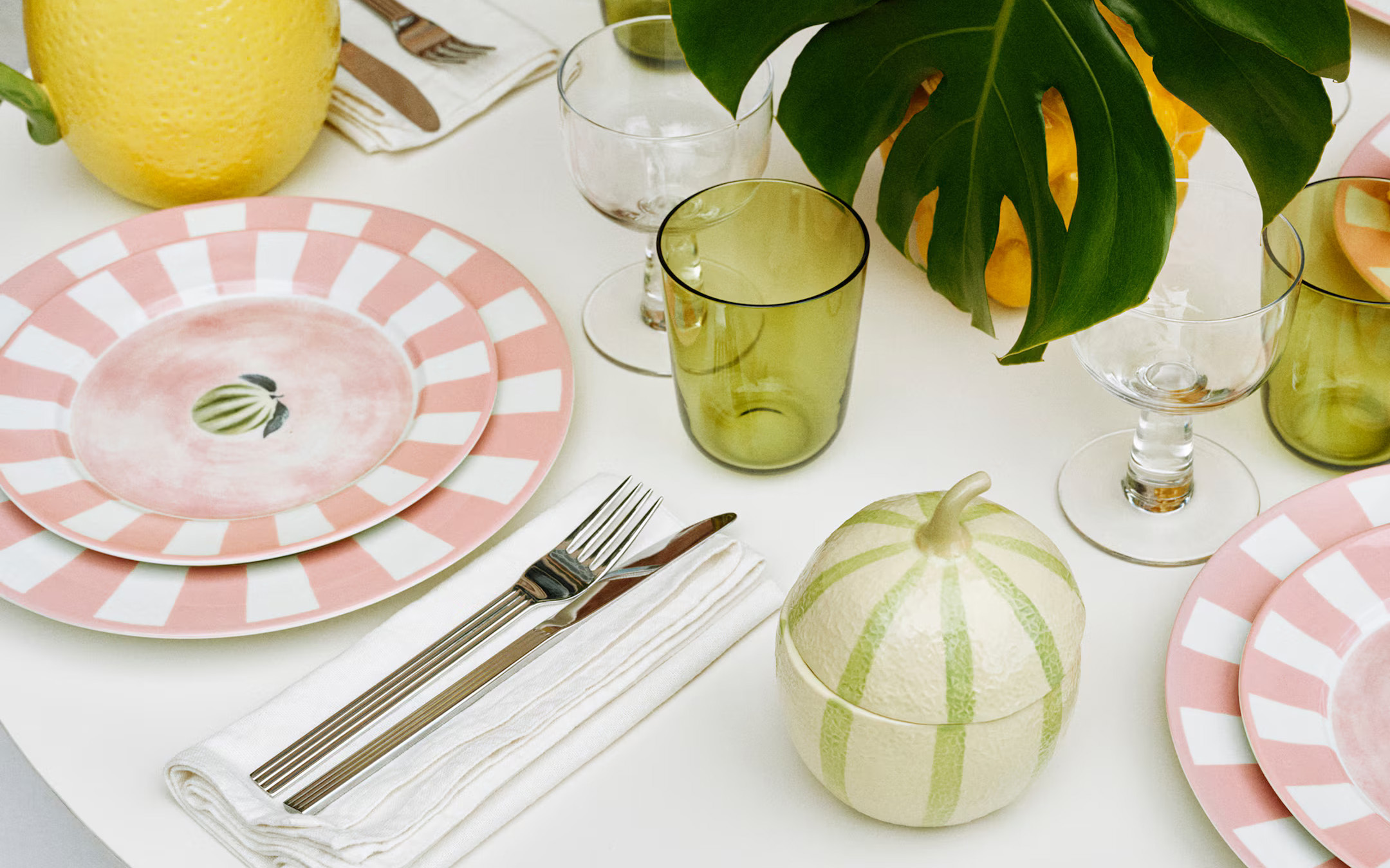 a summer table with a white tablecloth, pink and white stripe plates and a storage jar in the shape of a melon