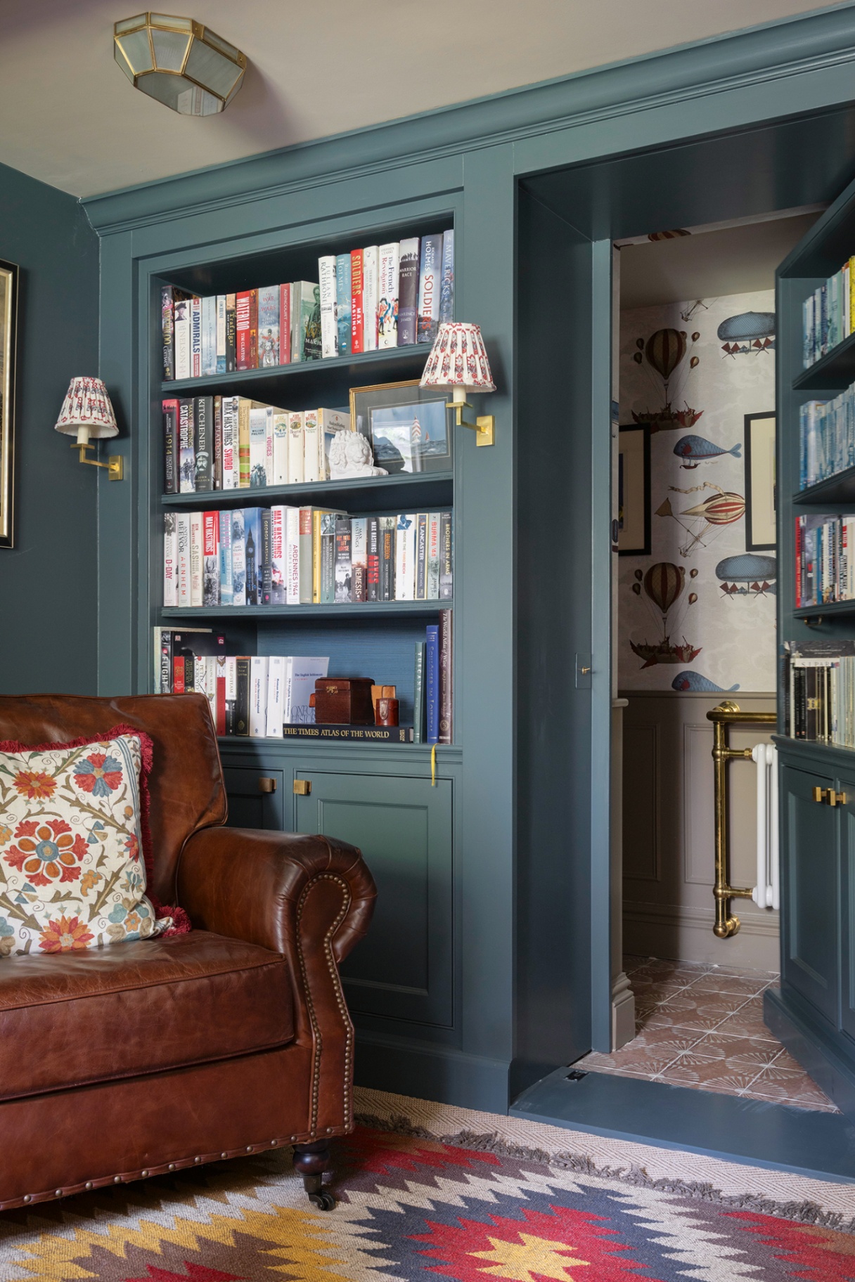 A home office with built-in bookcases painted in dark blue, which are filled with books and wall lights. A leather armchair and patterned rug are in the foreground. To the right, a jib door is open to reveal a hidden cloakroom