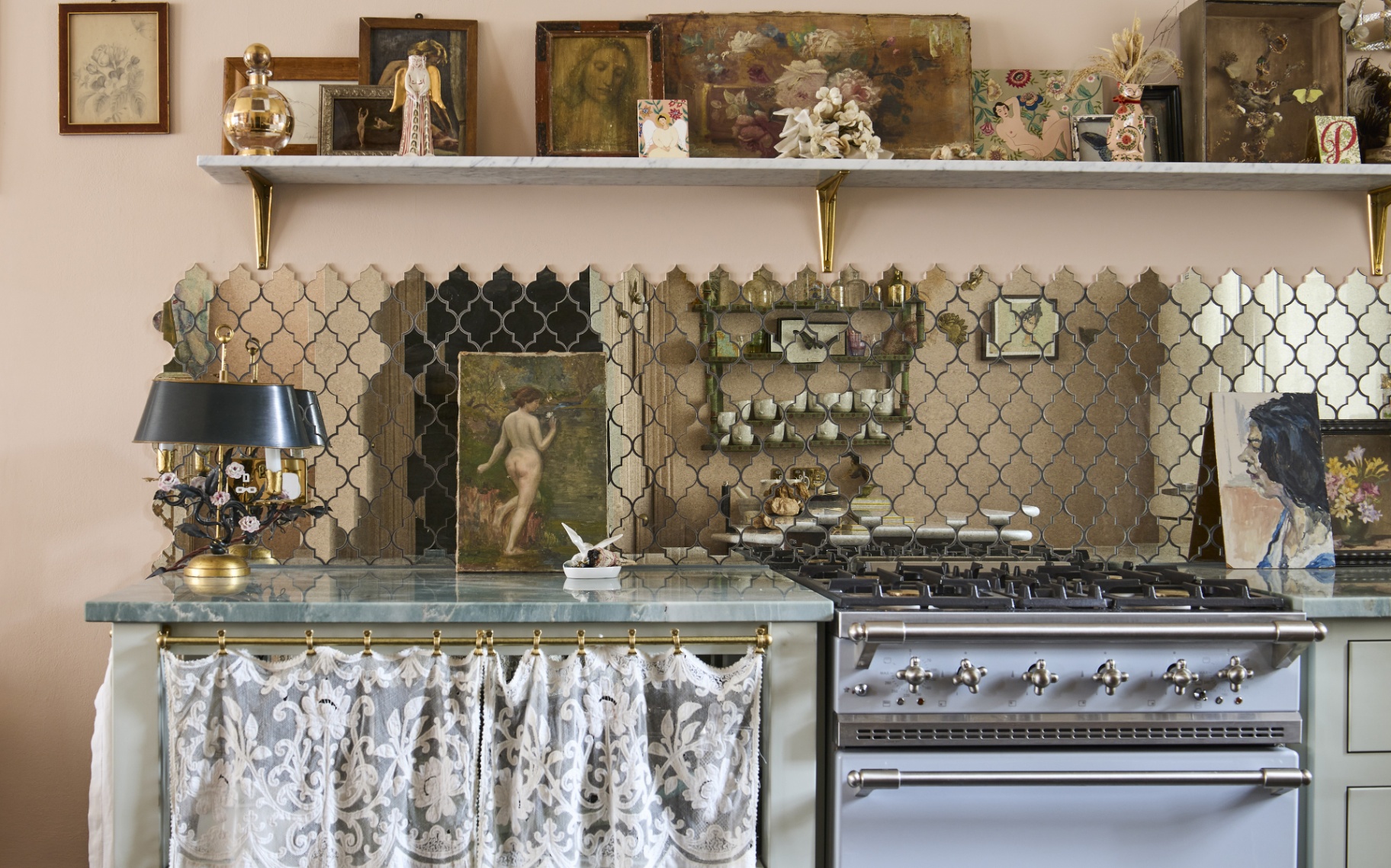 a kitchen corner wtih a pale blue range cooker, a cabinet with a fabric skirt, a long mirrored tile splashback and a shelf above with antique art and decor