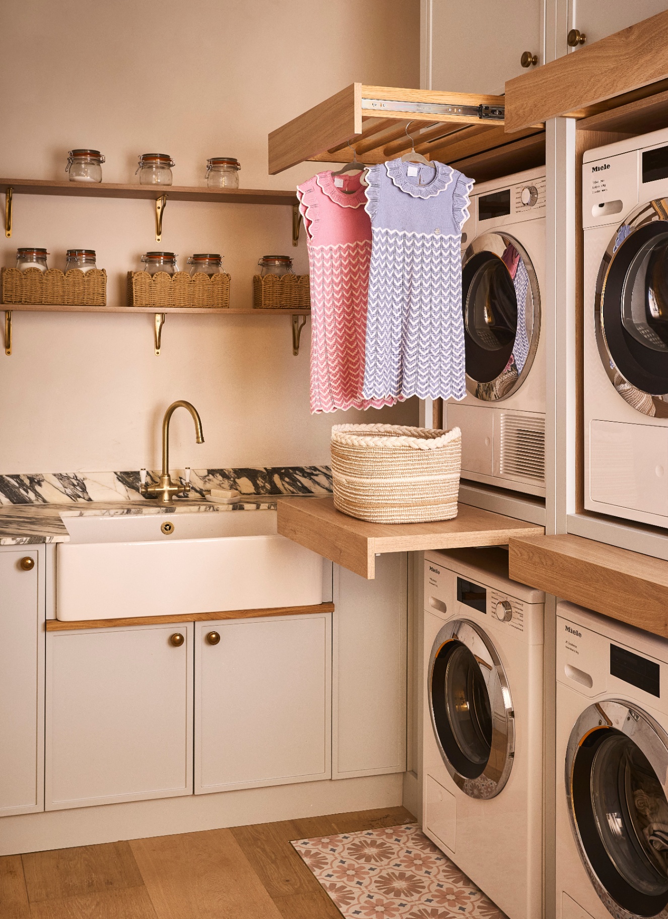 a utility room with cream cabinets, white and black marble countertops, a belfast sink, 4 stacked washer and dryers and open shelving with wicker baskets
