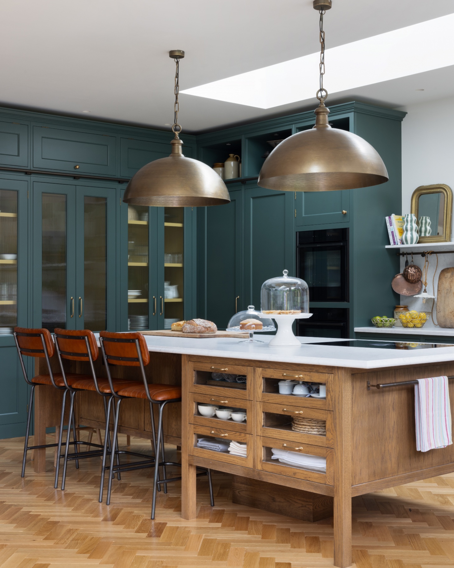 a kitchen with teal blue cabinetry, 2 large oversized dome pendants hanging above a wooden kitchen island with a white marble top and 3 brown leather chairs tucked under it