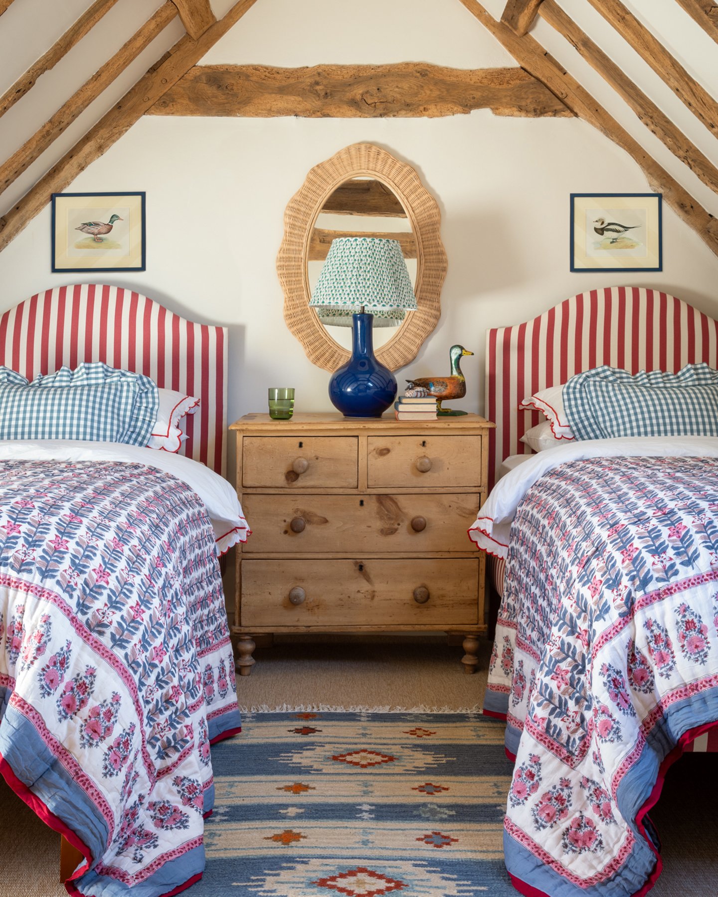 an eaves bedroom with wooden beams, two single beds with a white and red stripe headboard and a blue, white, pink and red floral bedspread