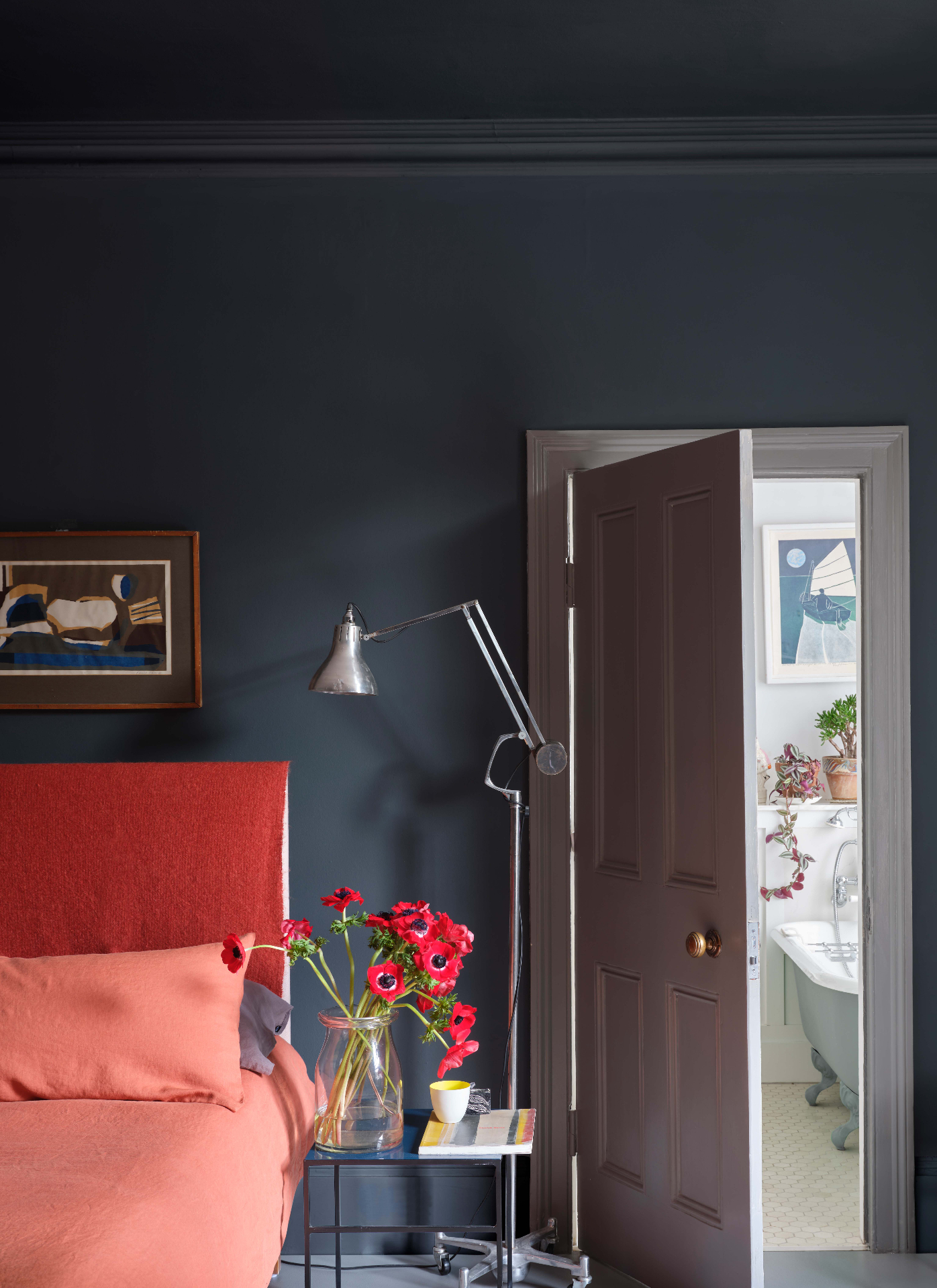 Dark slate blue bedroom with color-drenched ceiling and walls, an orange padded headboard, peach bedspread, tall silver floor lamp, and red flowers on a nightstand