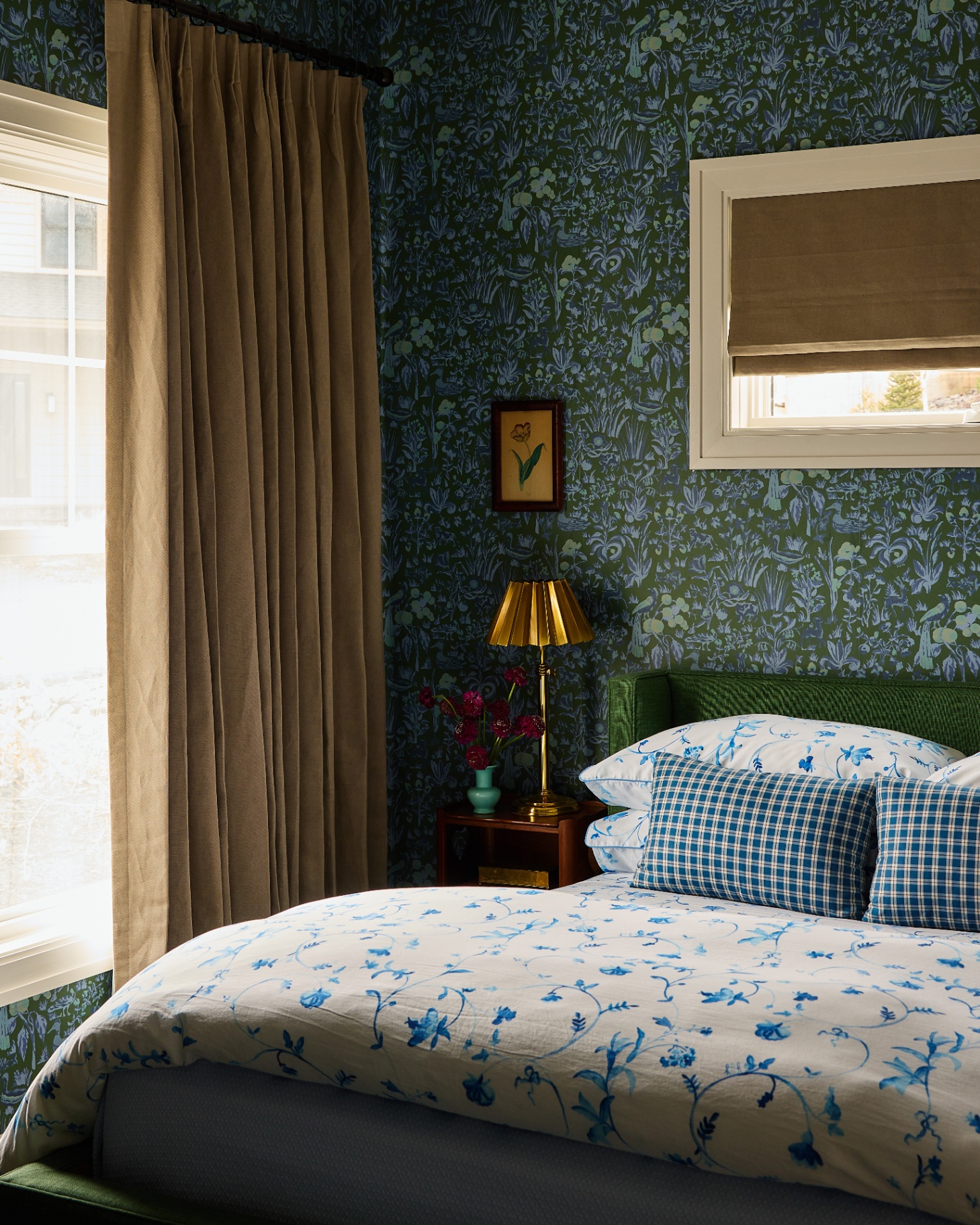 a bedroom with dark green and blue floral wallpaper, ochre curtains and blind,a bed with white and blue floral bedding and a gold bedside lamp