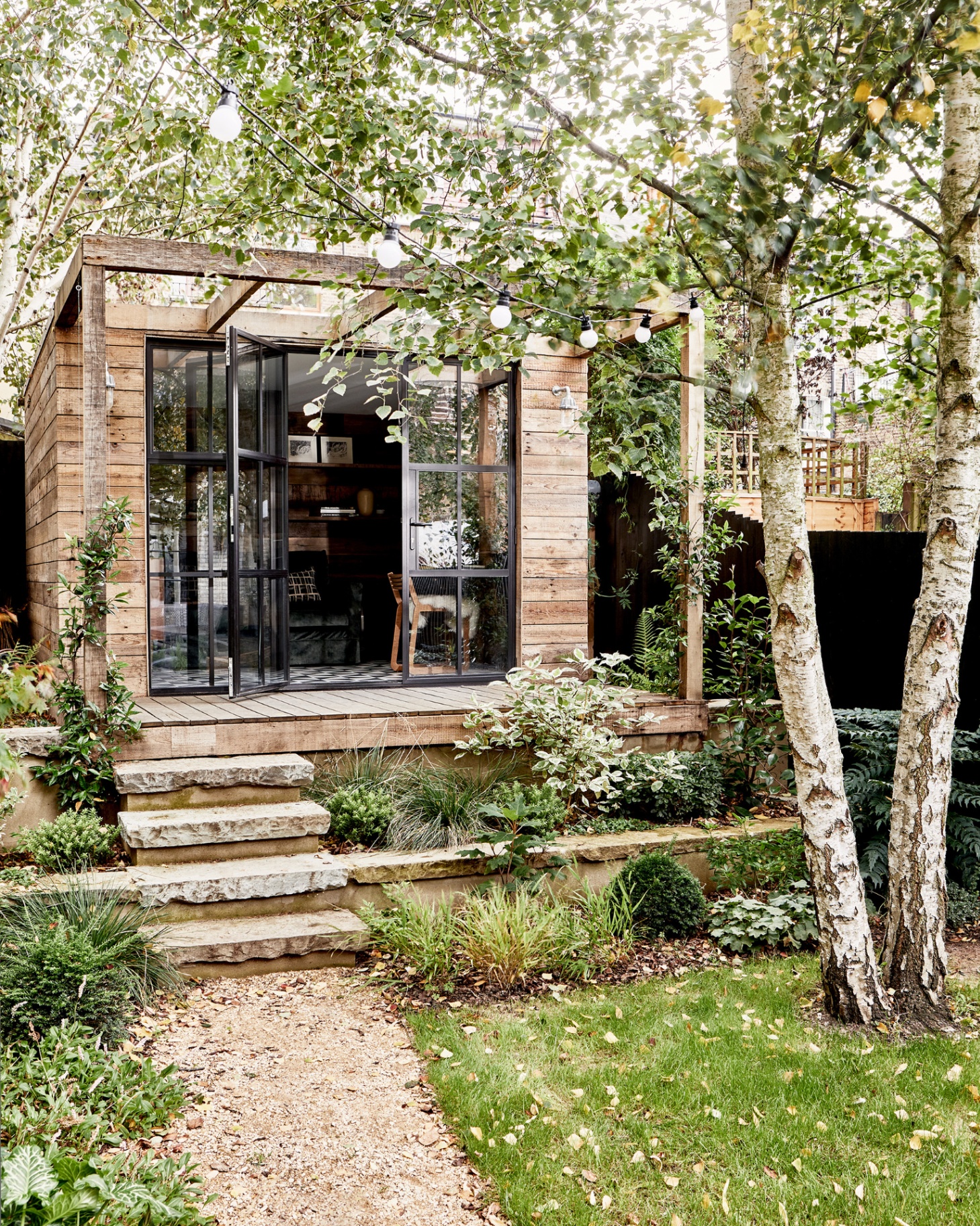 a wooden garden room with black glass doors with steps leading down to a gravel path and festoon lights hanging from the trees