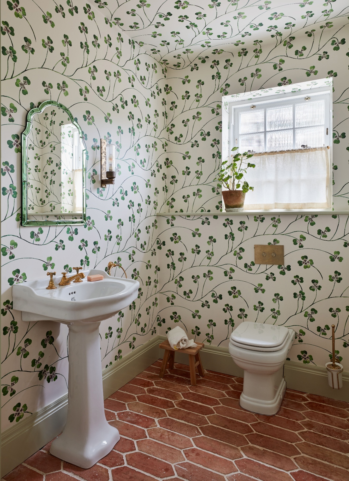 a downstairs bathroom with brick tiled flooring, a white toilet and sink, a white wallpaper with green trailing vines, an art deco green arch mirror and a window with a sheer cafe curtain