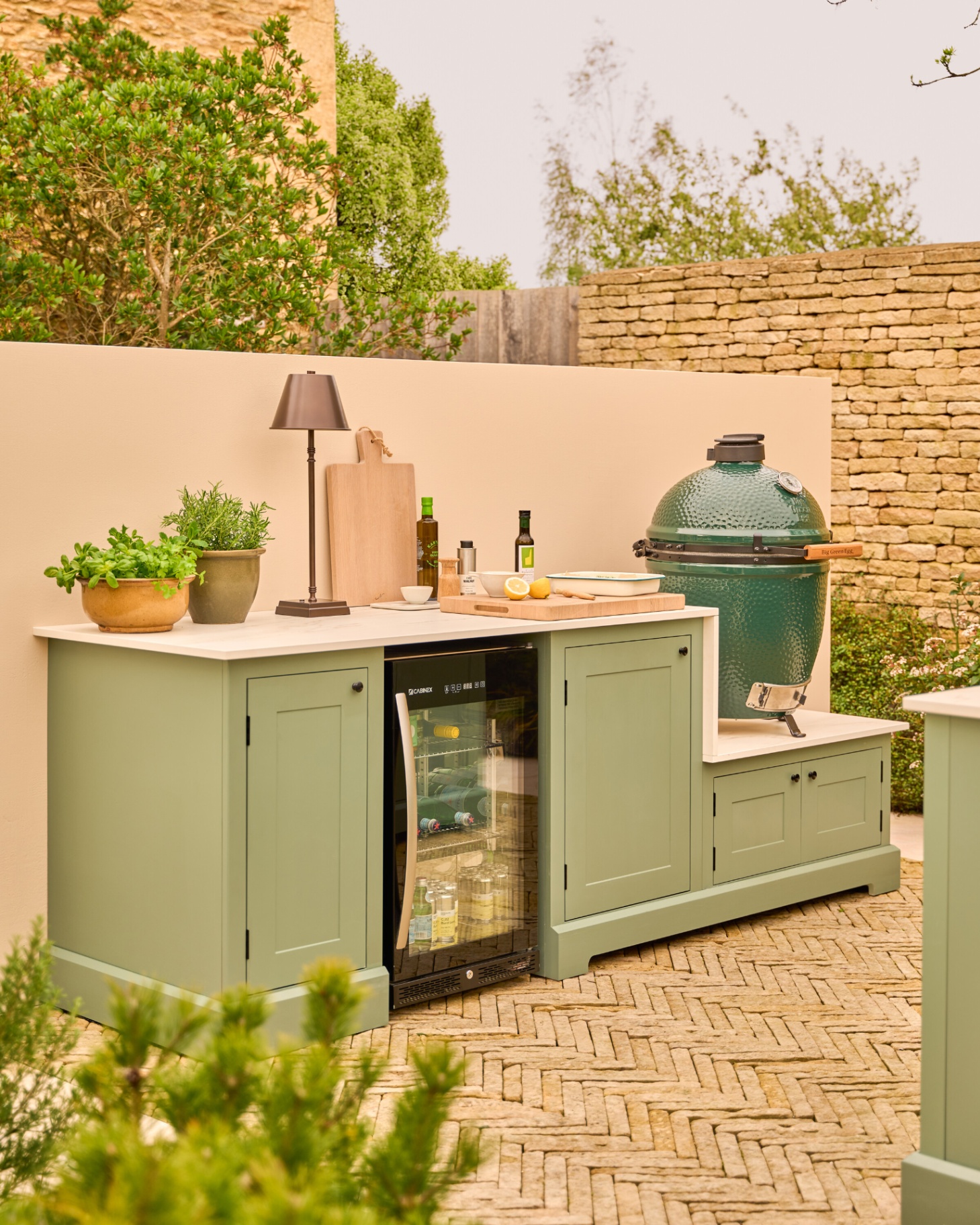 a sage green outdoor kitchen with a big green egg ceramic bbq, an outdoor fridge and a portable lamp