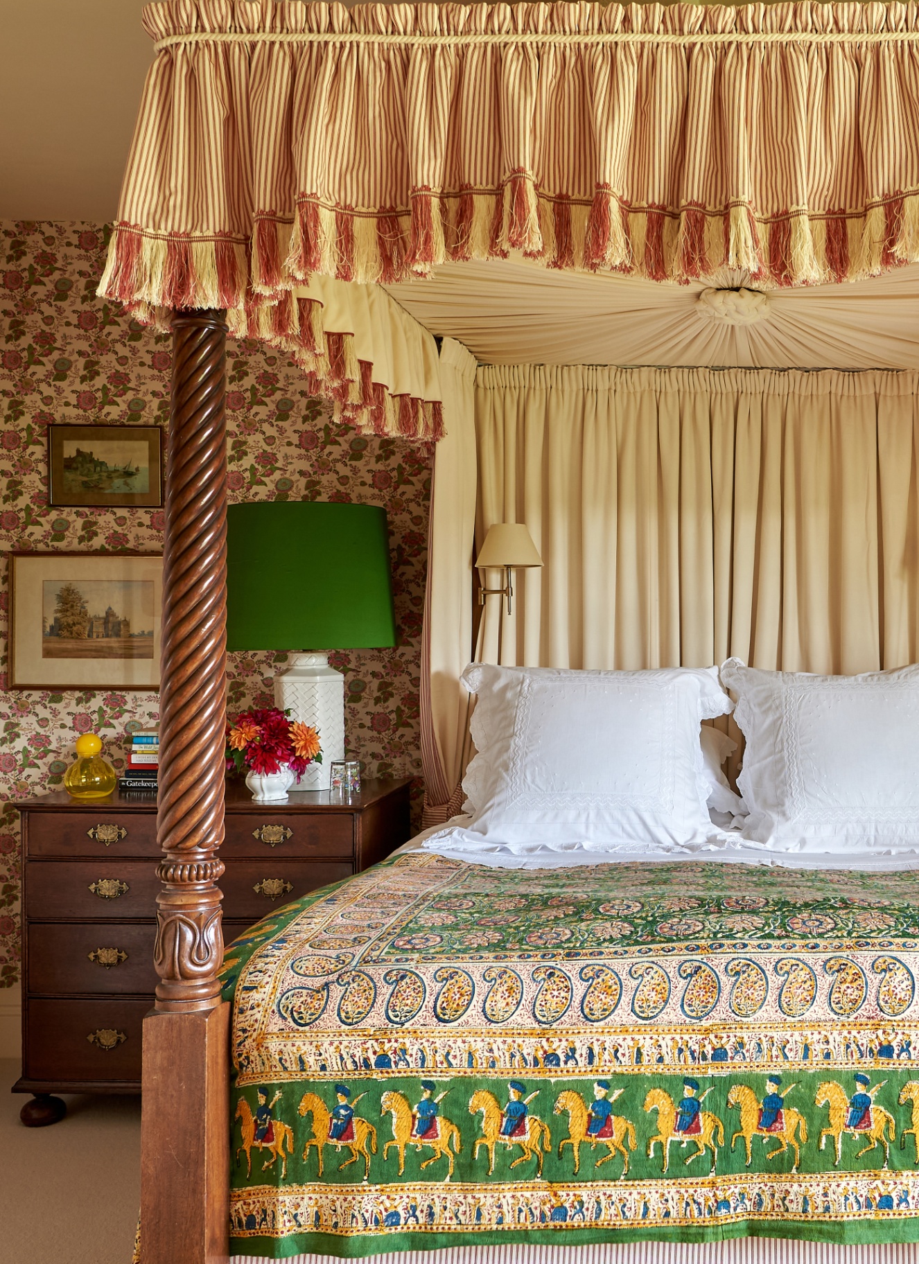 Opulent four-poster bed with a fringed canopy, striped valance, white crisp linens, and an ornate green paisley bedspread, set against floral wallpaper and a wooden chest of drawers topped with a bold green lamp