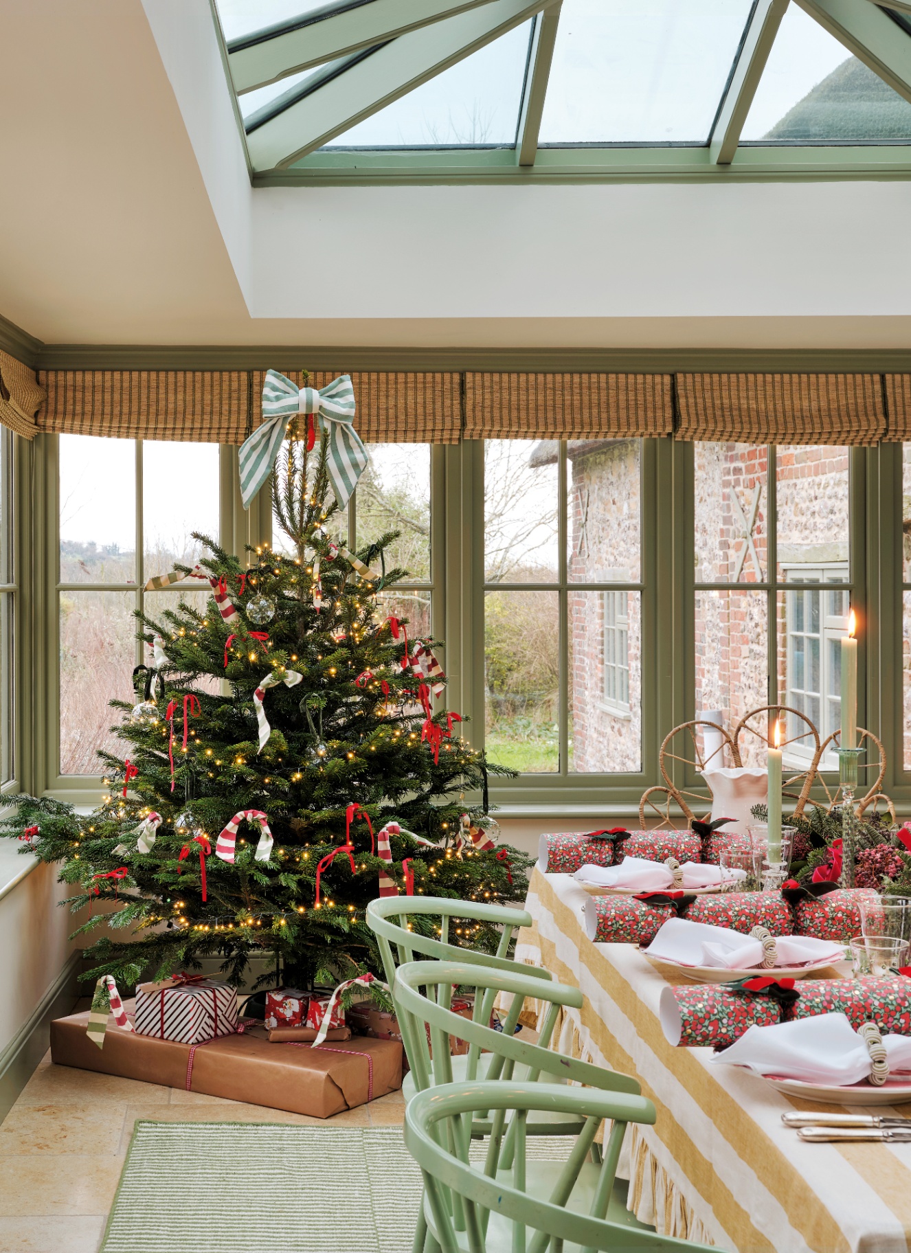 A Christmas tree topped with a large striped fabric bow standing in a sunlit garden dining room near a decorated table